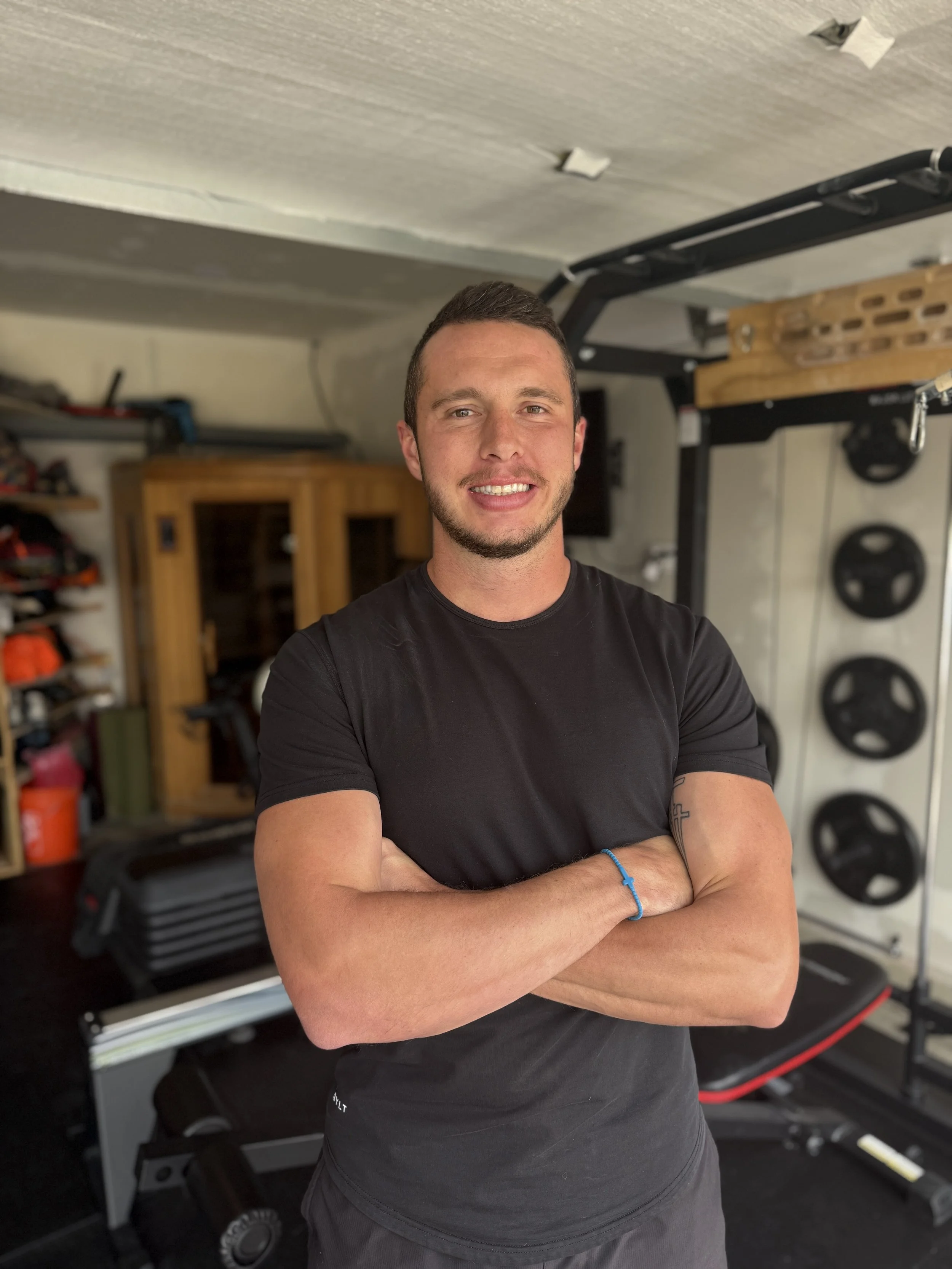 A young man with short brown hair and a beard, wearing a black t-shirt, standing with arms crossed in a home gym. In the background, there are weight plates, a workout bench, and a wooden cabinet.