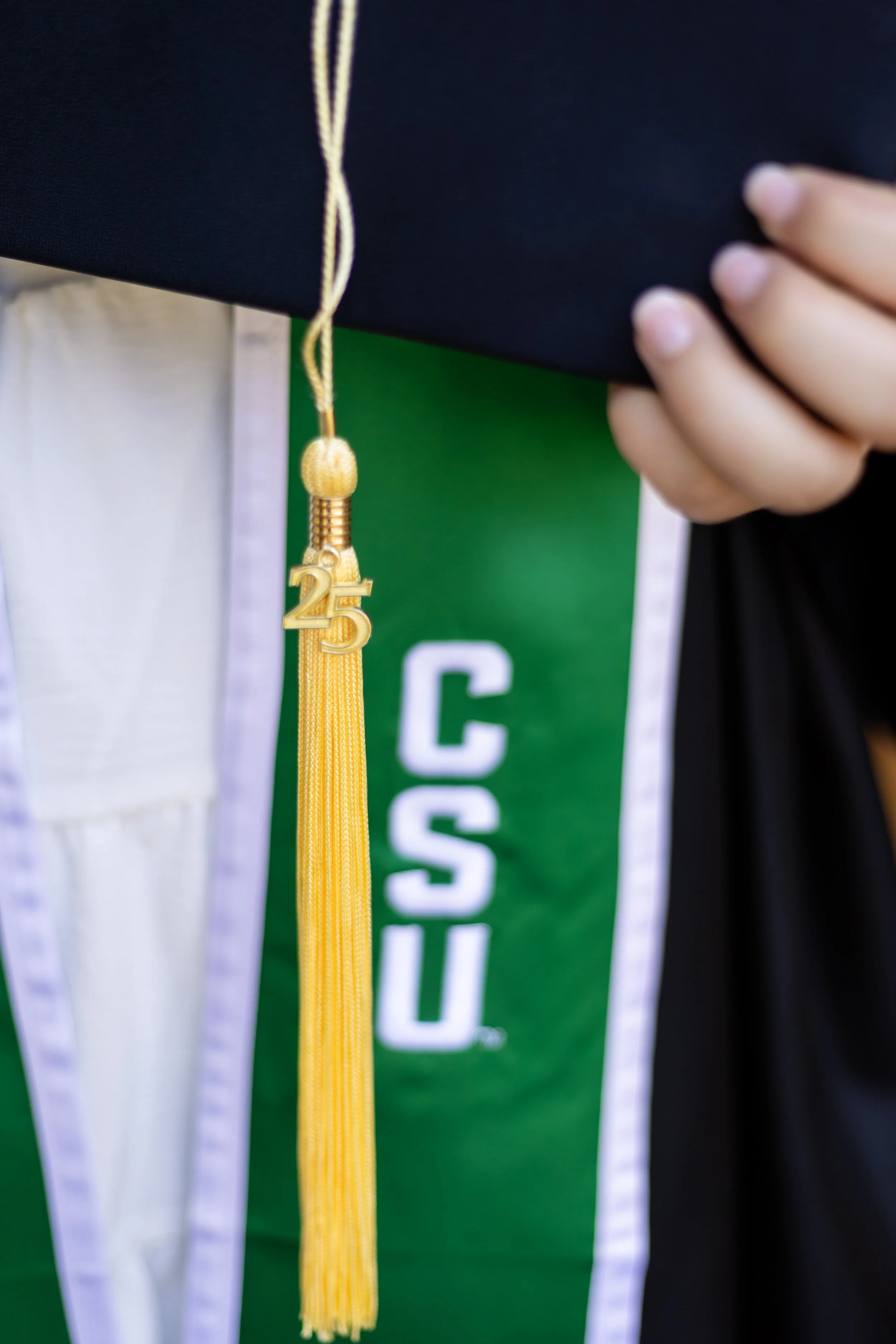 Close-up of a person's hand holding a black diploma cover with a gold tassel hanging from it. The tassel has a charm with the number 25.