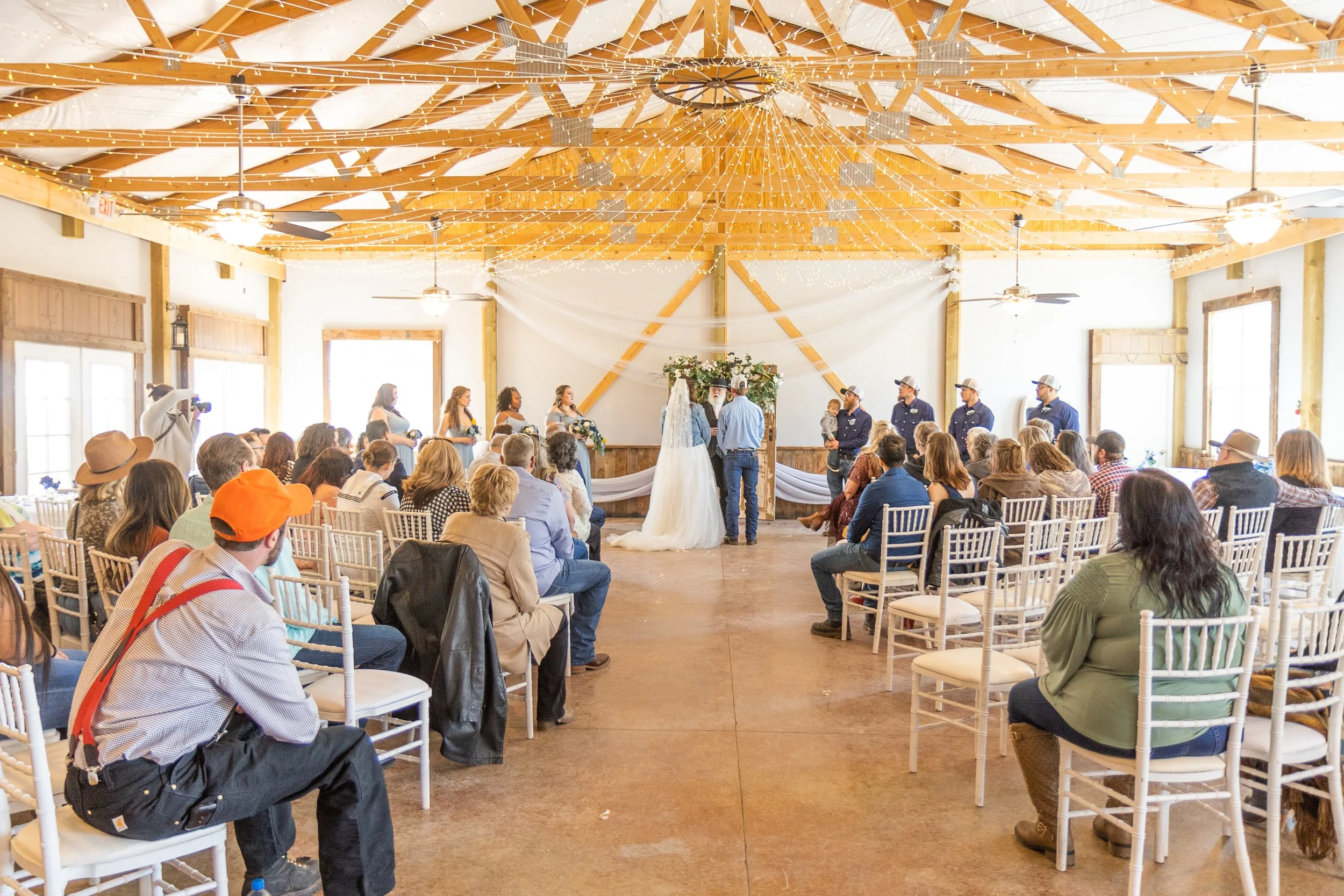Indoor wedding ceremony with guests seated on white chairs, officiant and bridesmaids standing in front of the bride and groom, all under a wooden ceiling decorated with string lights. Once Upon An Event Center, Kiowa, CO