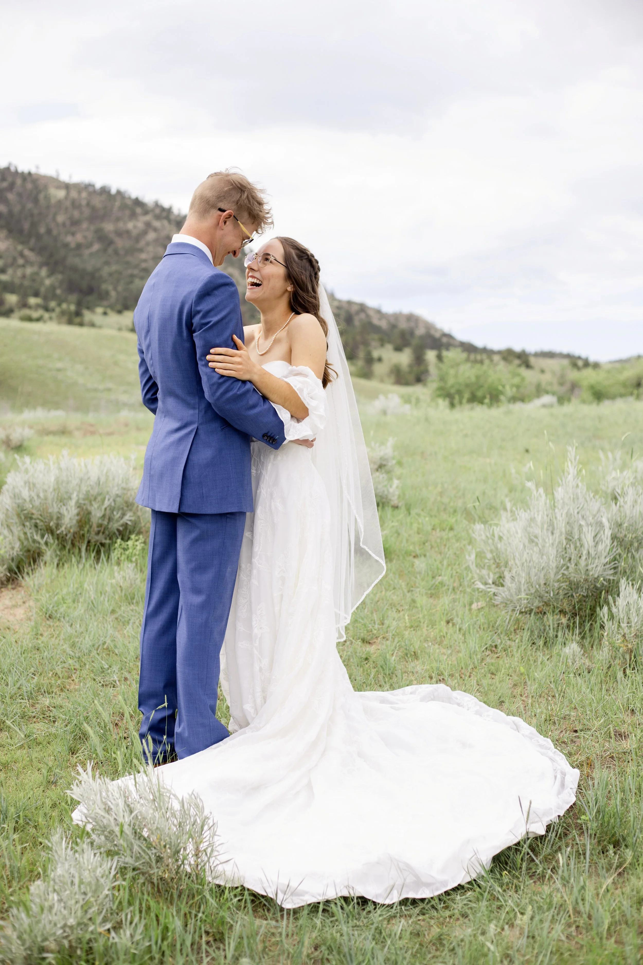 A newlywed couple standing in a grassy field, embracing and smiling at each other on their wedding day.