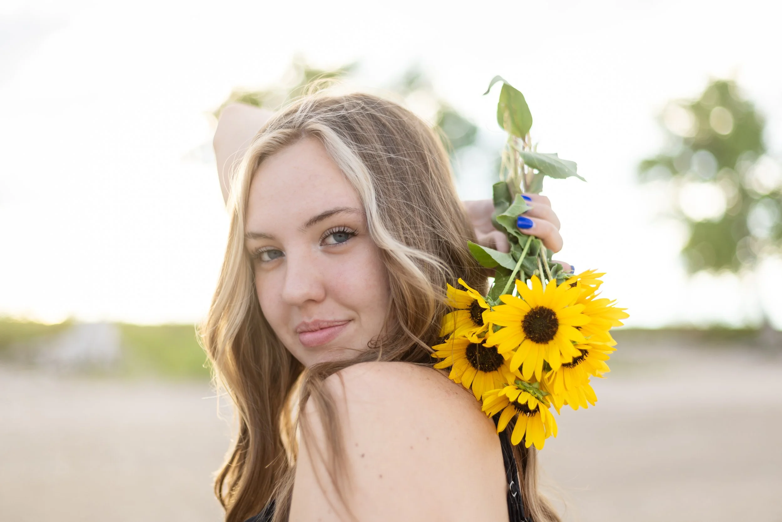 A woman with long, wavy blonde hair holds a bouquet of bright yellow sunflowers on her shoulder, outdoors with soft sunlight and blurred greenery in the background.