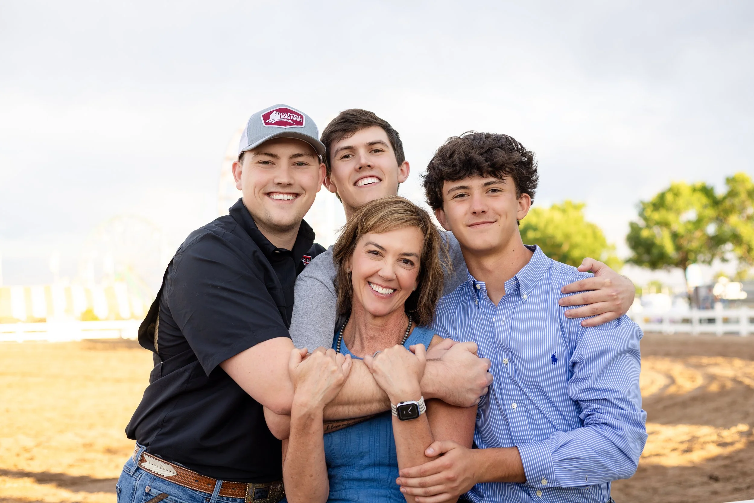A group of five smiling people, including three young men and a woman, embracing outdoors in a park with trees and a Ferris wheel in the background.