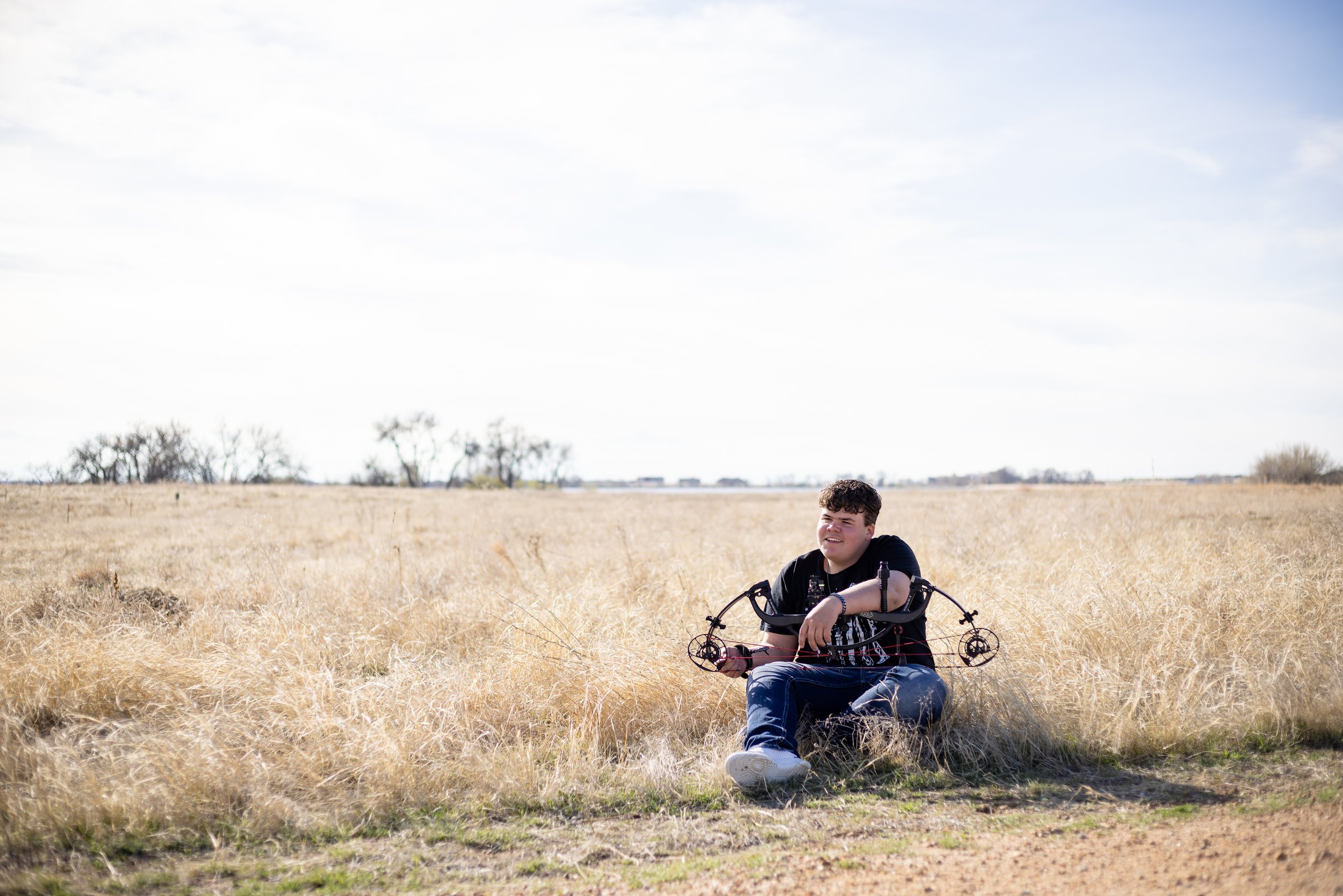 Young man sitting on the grass in a wide open field, smiling, with some trees in the background and a cloudy sky overhead.