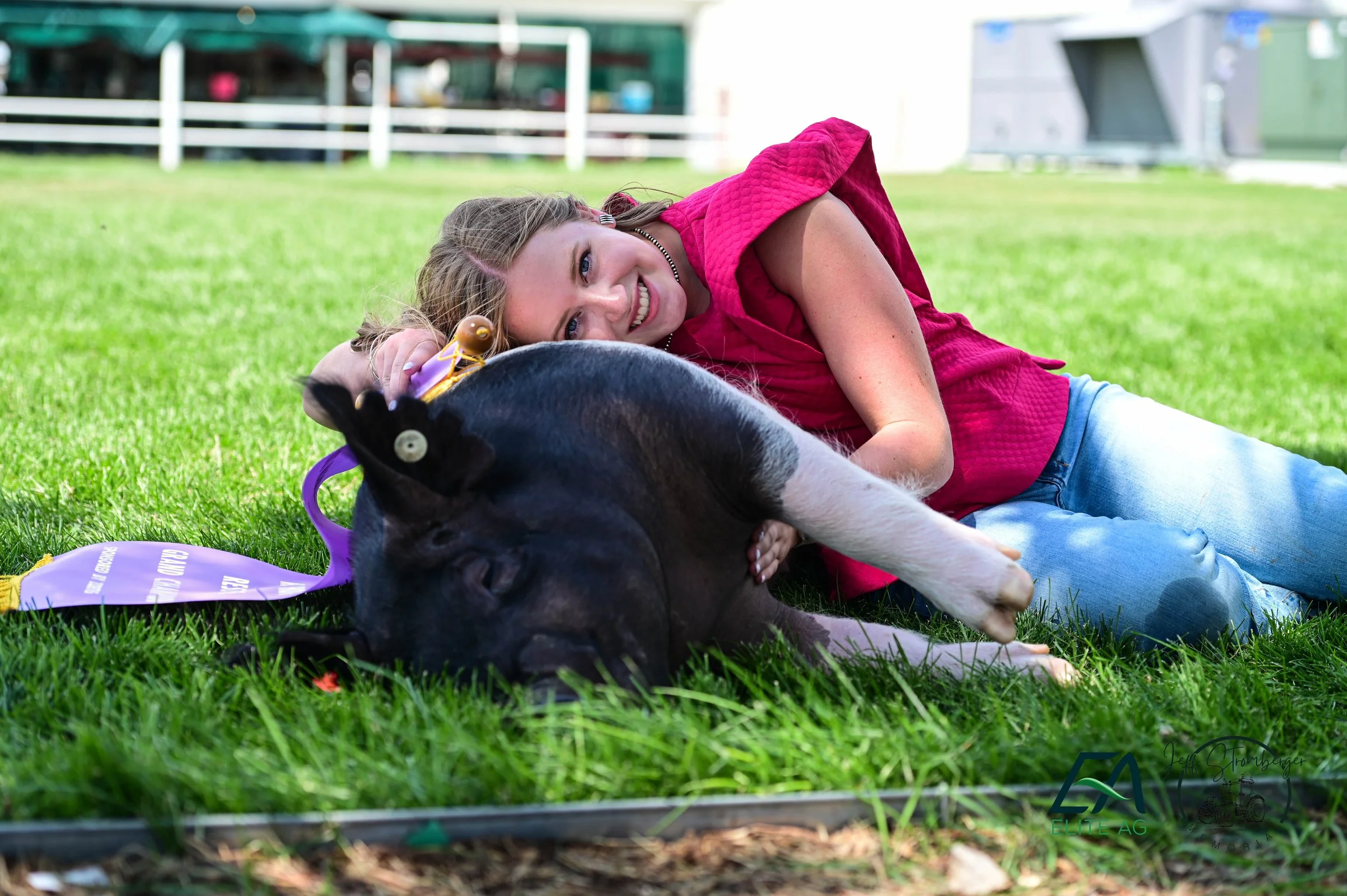 A young woman with blonde hair and blue eyes lying on green grass alongside a black and white puppy with a yellow ribbon around its neck. The woman is smiling and resting her head on the puppy, in an outdoor setting with a white fence and buildings in the background