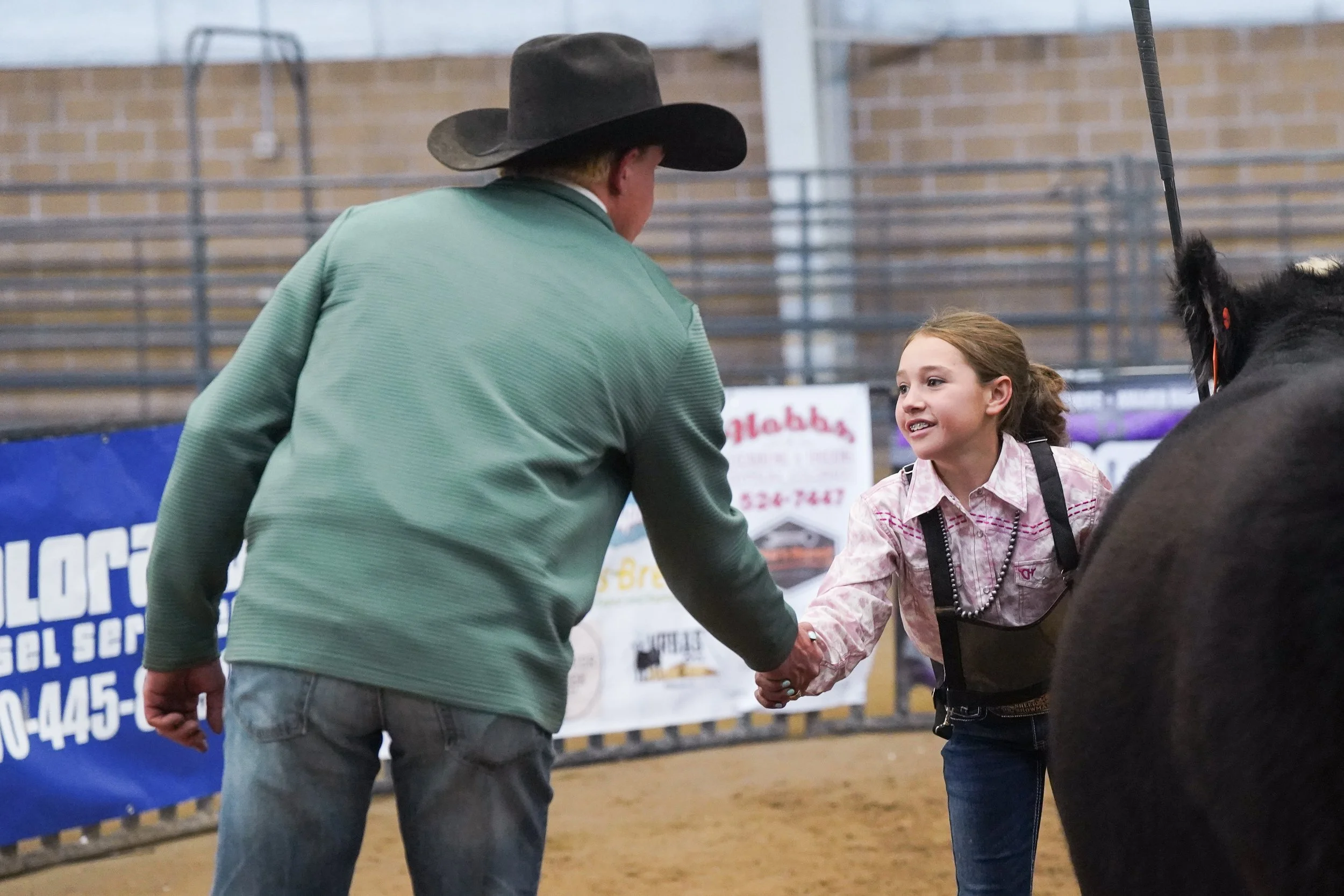 A man and a young girl shake hands inside a rodeo arena. The man wears a large black cowboy hat and a greenish jacket, while the girl wears a pink shirt with a black vest and a pearl necklace. A horse's head is partially visible on the right. The background has brick walls and signage related to rodeo events.