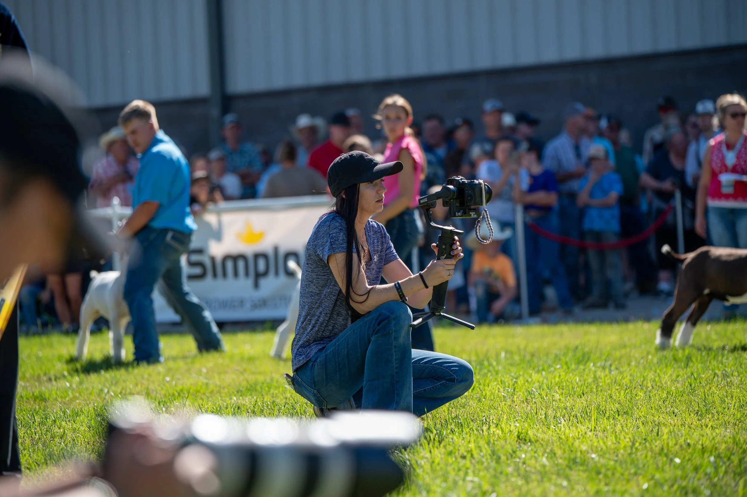 A woman kneeling on grass holding a camera stabilizer during an outdoor event, with a crowd of spectators behind her, some wearing hats and sunglasses, and a woman with a pink shirt and glasses in the background.