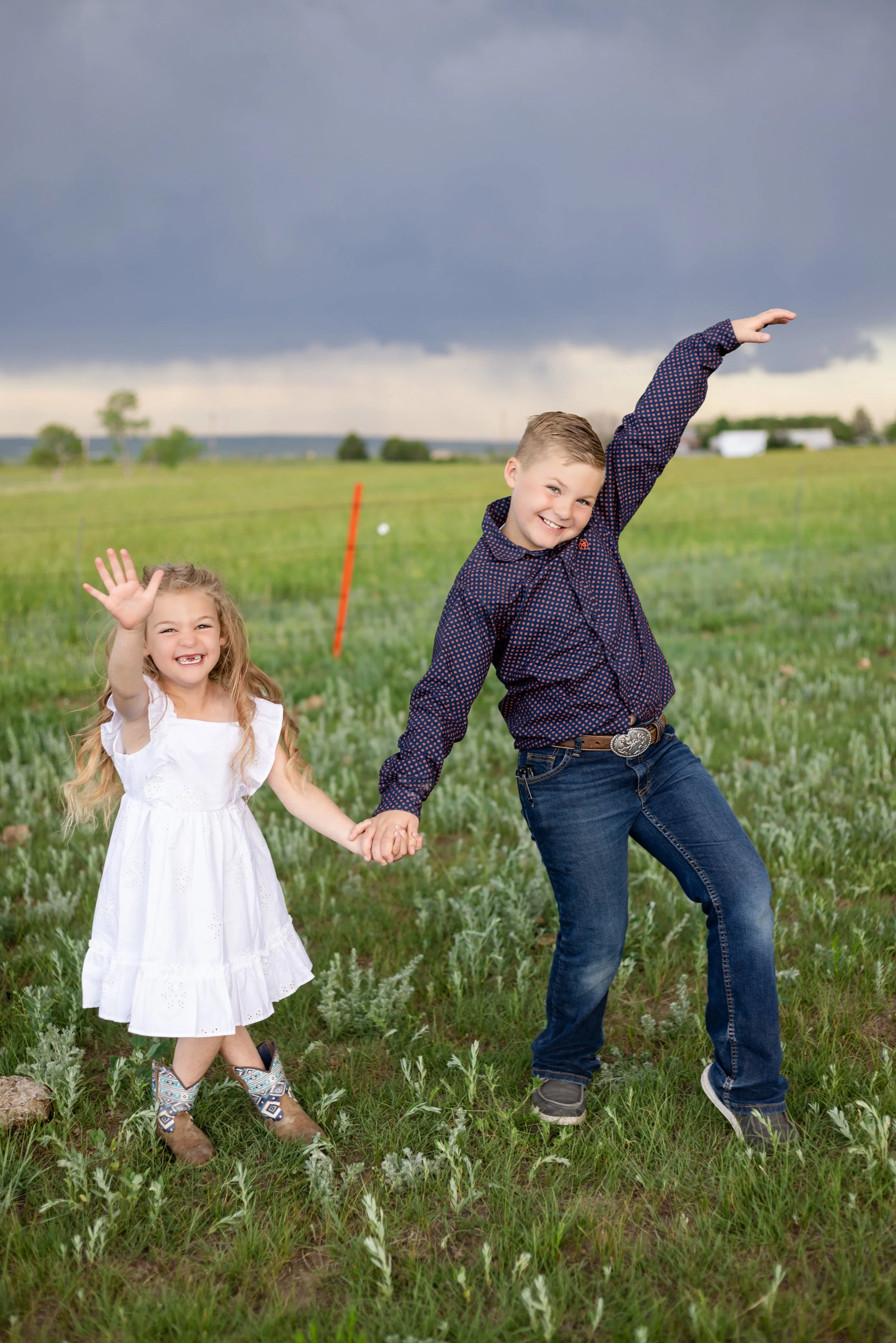 A young girl and a boy holding hands in a field with green grass and plants, smiling and waving, under a dark cloudy sky.