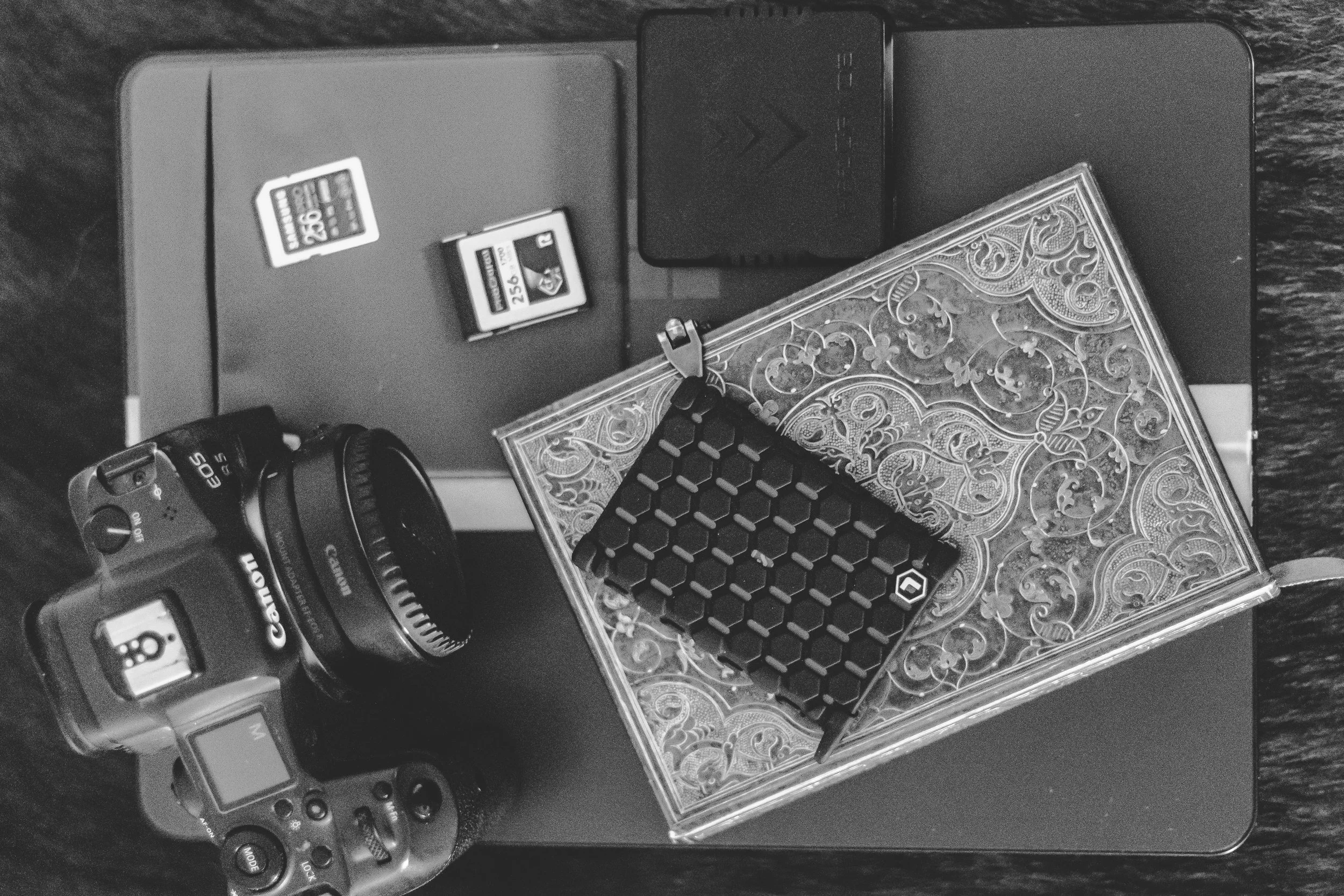 Black and white photograph of a camera, two SD cards, a decorative card box, and electronic accessories on a dark textured surface. Beyond the Ring Education