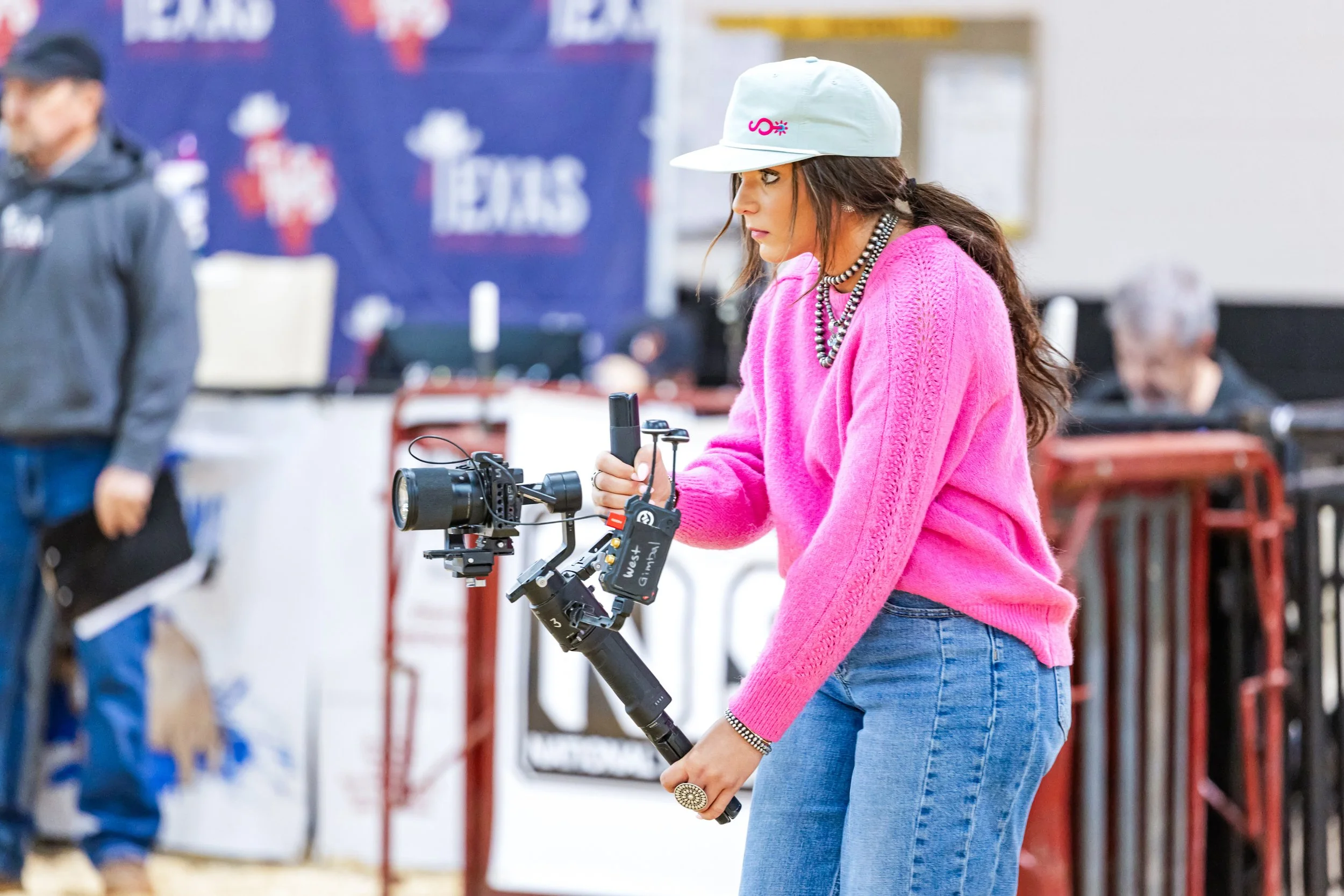 A woman wearing a white cap, pink sweater, and jeans operating a camera mounted on a stabilizer at an indoor event.