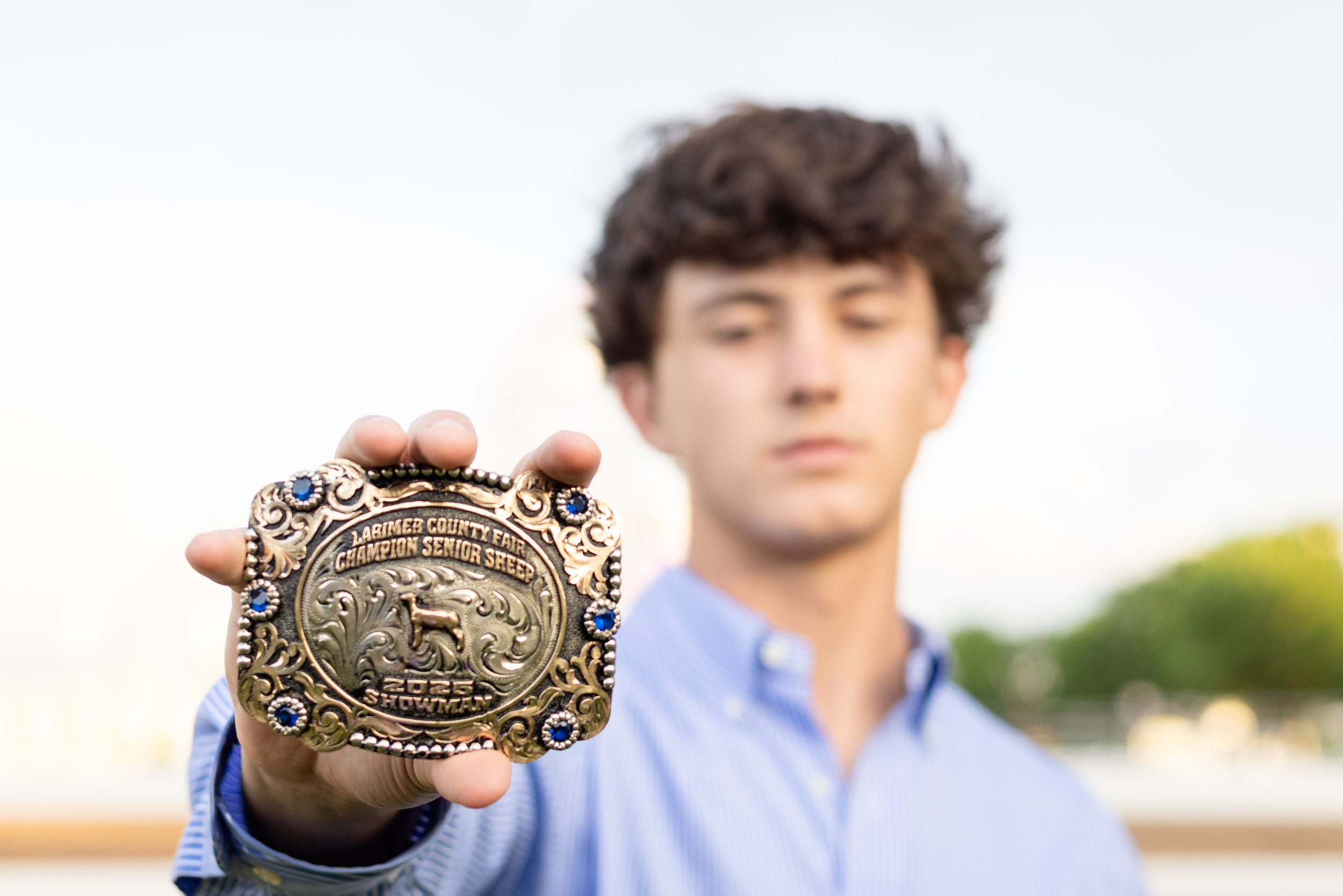 Young man holding up a gold and black championship belt with blue accents, out of focus background with sky and greenery.