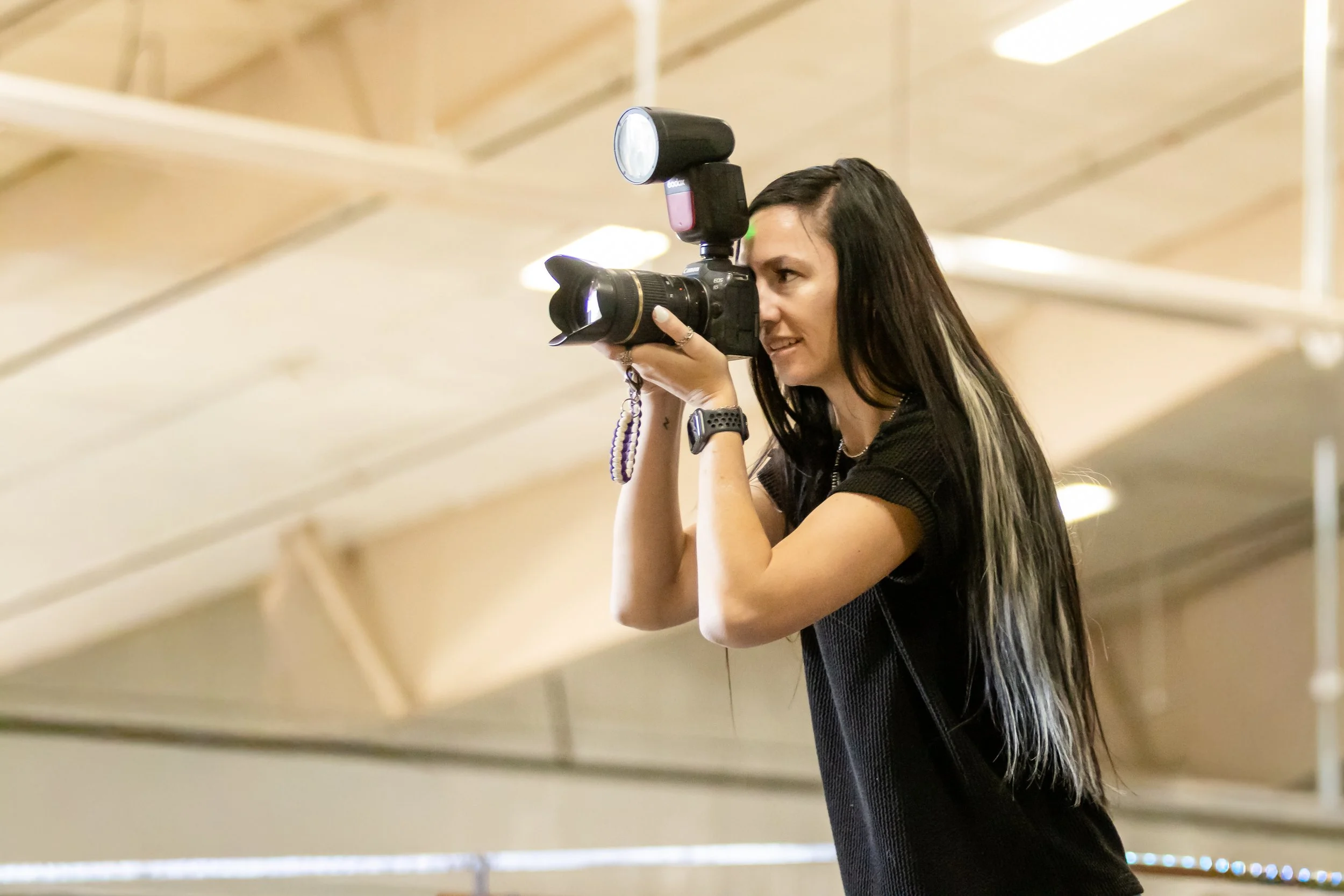 Woman with long dark hair taking a photograph indoors using a camera with a flash attachment.