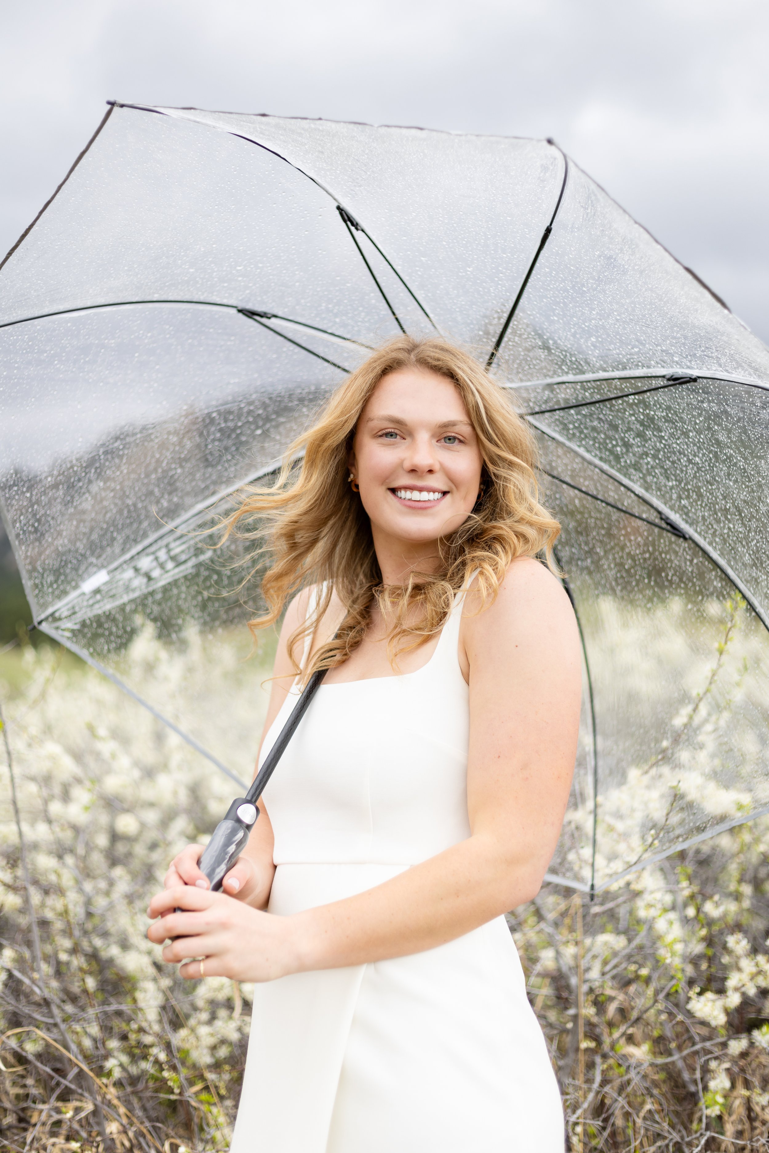 A smiling woman in a white dress holding a transparent umbrella with raindrops, outdoors amidst blossoming bushes.
