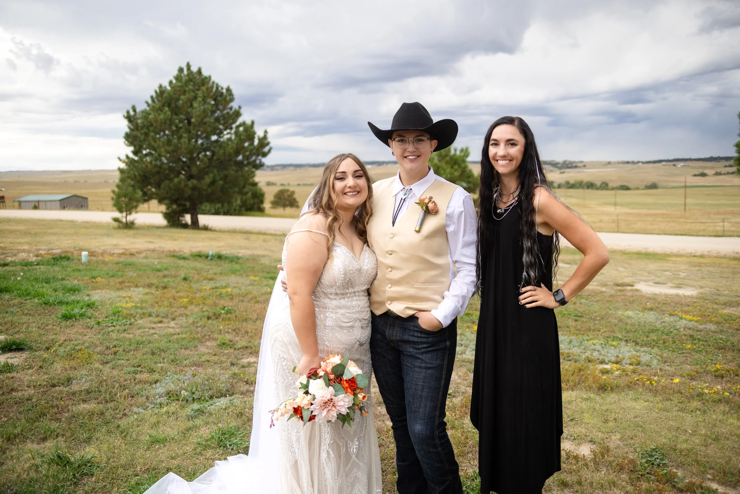 Three women standing outdoors in a grassy area, with open fields and trees in the background under a cloudy sky. The woman on the left is wearing a white lace wedding dress and holding a bouquet. The woman in the middle is dressed in cowboy attire with a hat, vest, and boutonniere. The woman on the right is wearing a black dress with layered necklaces