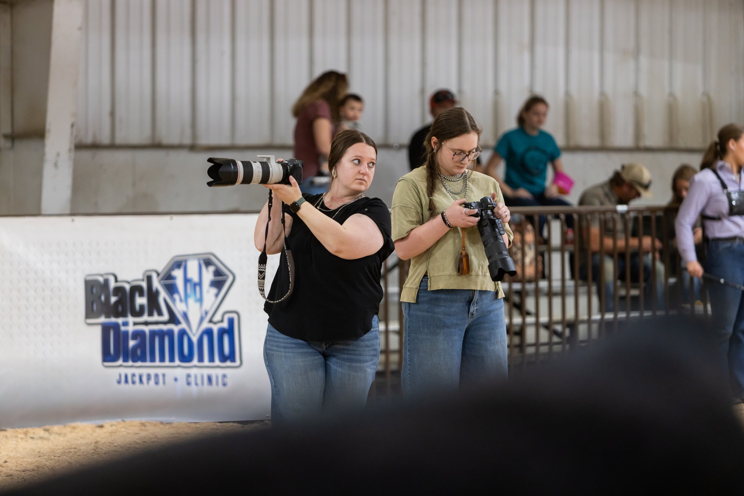 Two women standing in an indoor arena, one holding a camera with a large telephoto lens and the other looking at her phone. Other people are visible in the background watching or preparing for an event, with a sign that reads "Black Diamond Jackpot" partially visible.