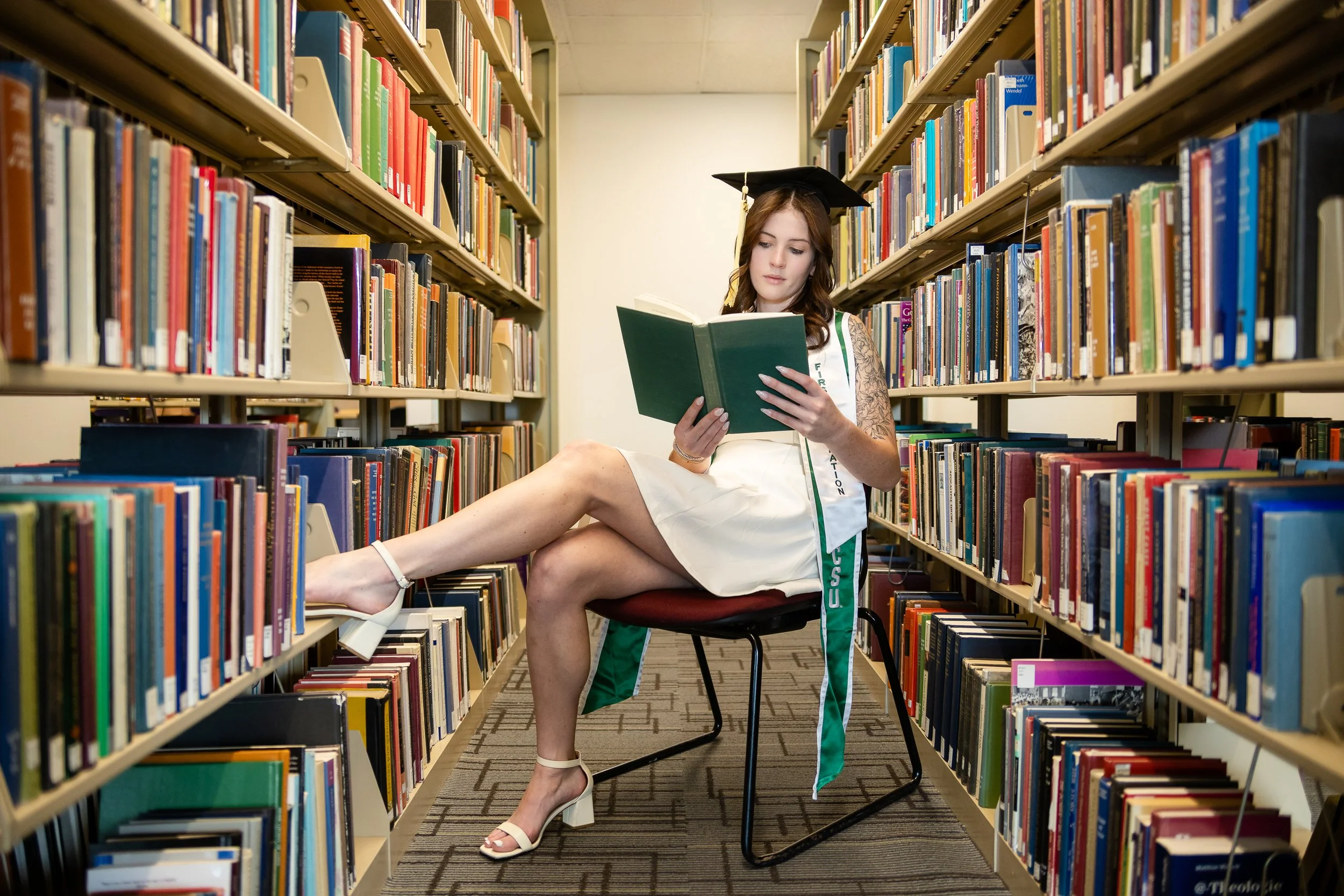 A young woman wearing a graduation cap and a white dress sitting on a chair between bookshelves in a library, reading a green book.