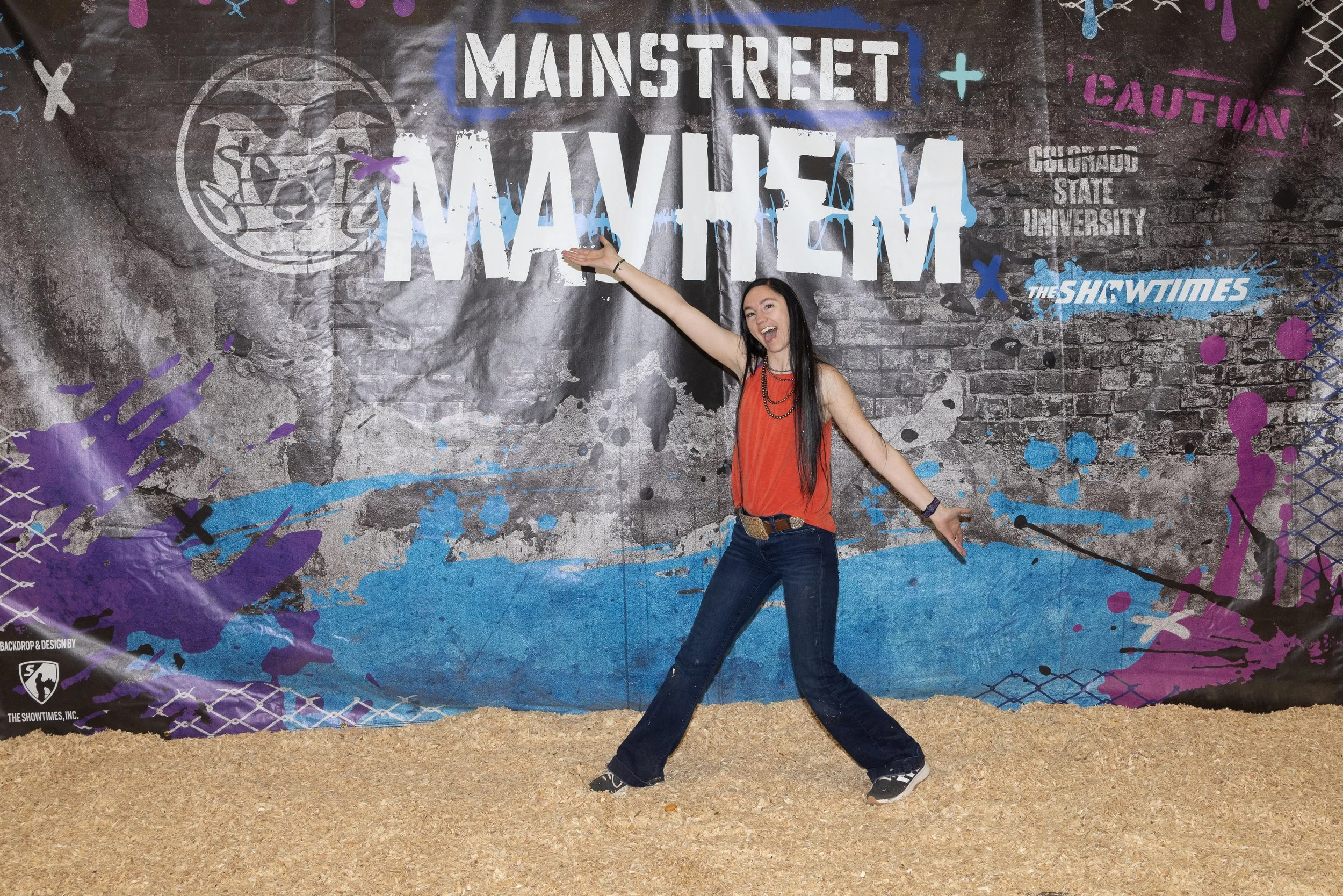 A woman with long black hair wearing an orange sleeveless top, dark jeans, and sneakers, standing with her arms outstretched and smiling in front of graffiti-style wall art that reads 'Main Street Memorial' and includes various colorful designs and logos.
