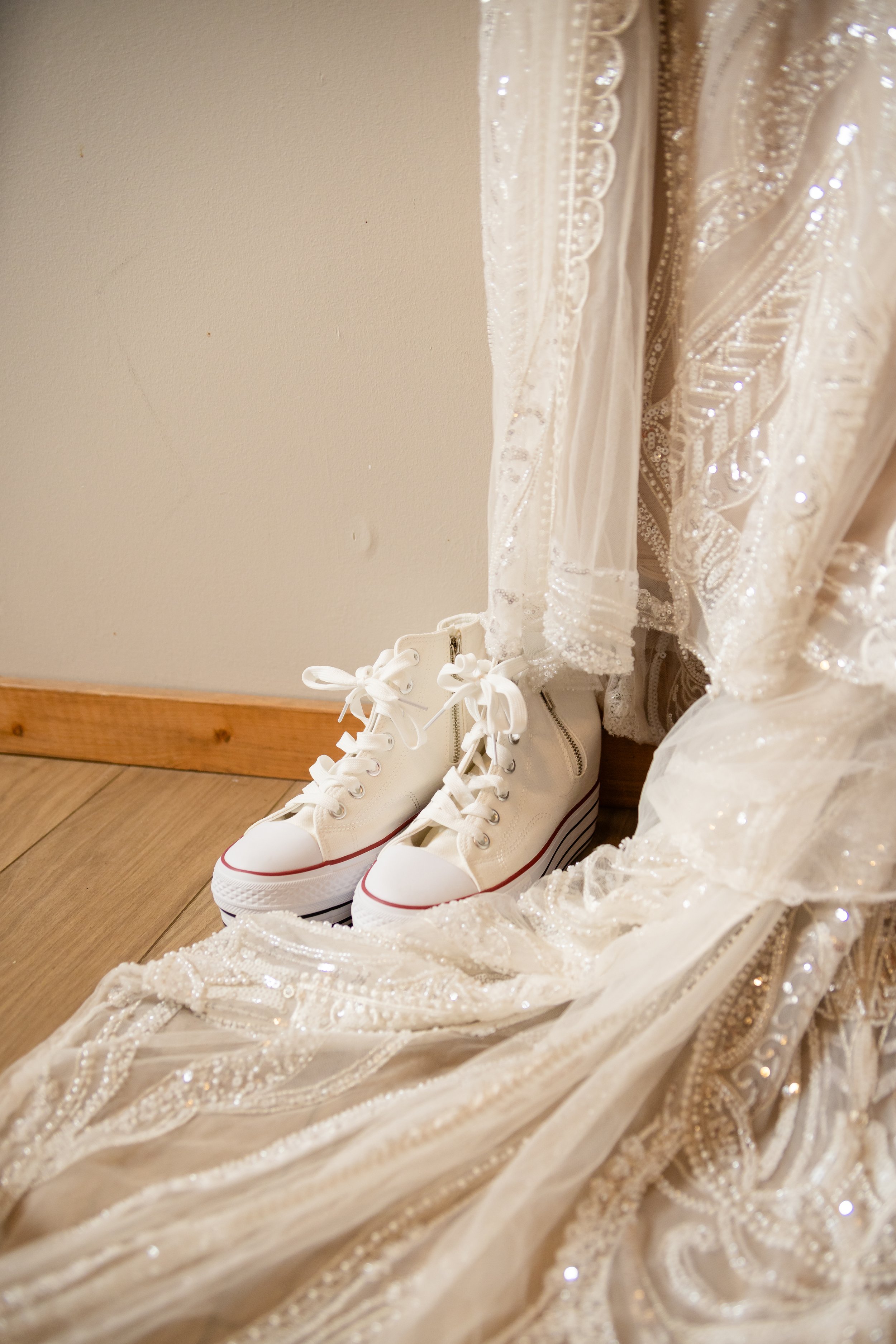 A pair of white high-top sneakers placed on a wooden floor beside a wedding dress with intricate beadwork and embroidery.