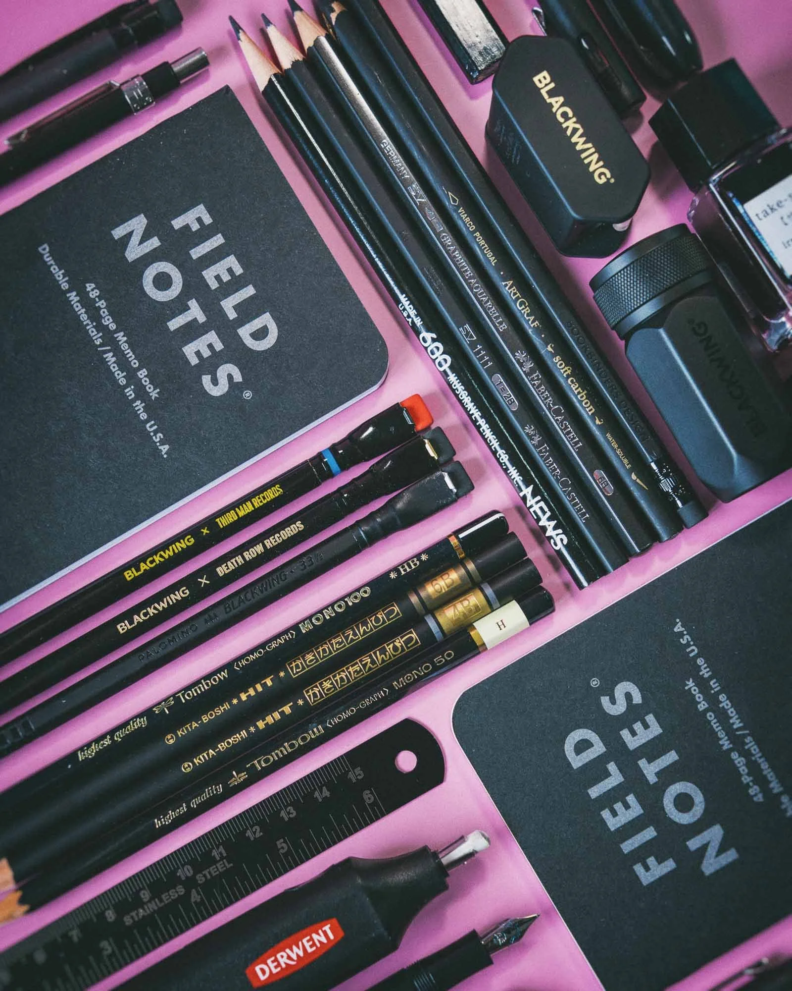 A flat lay of various black art supplies including pencils, pens, markers, a ruler, erasers, and a notebook on a pink background.