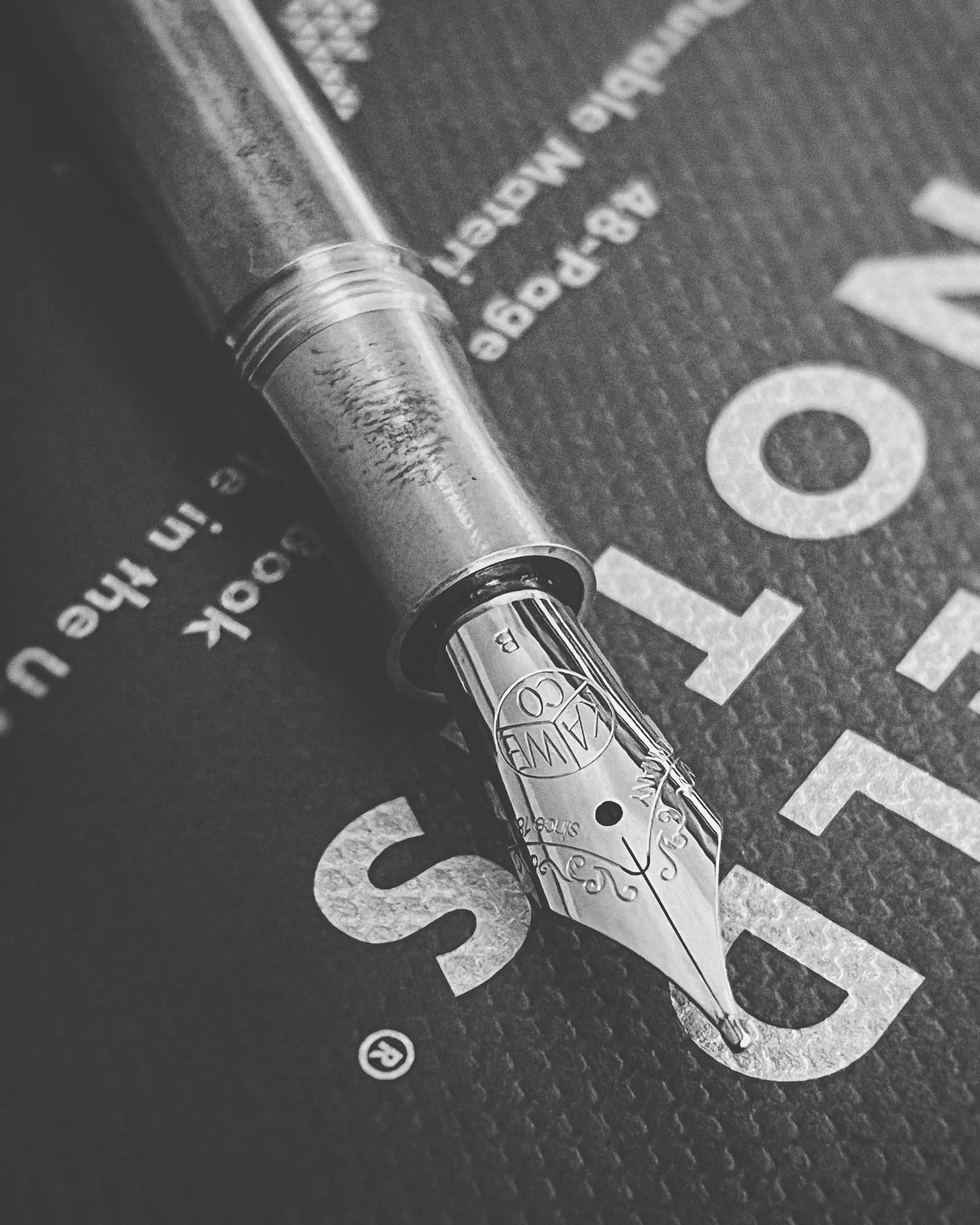 Close-up of a metallic fountain pen with intricate engravings on the nib, resting on a textured background that contains partially visible printed text.
