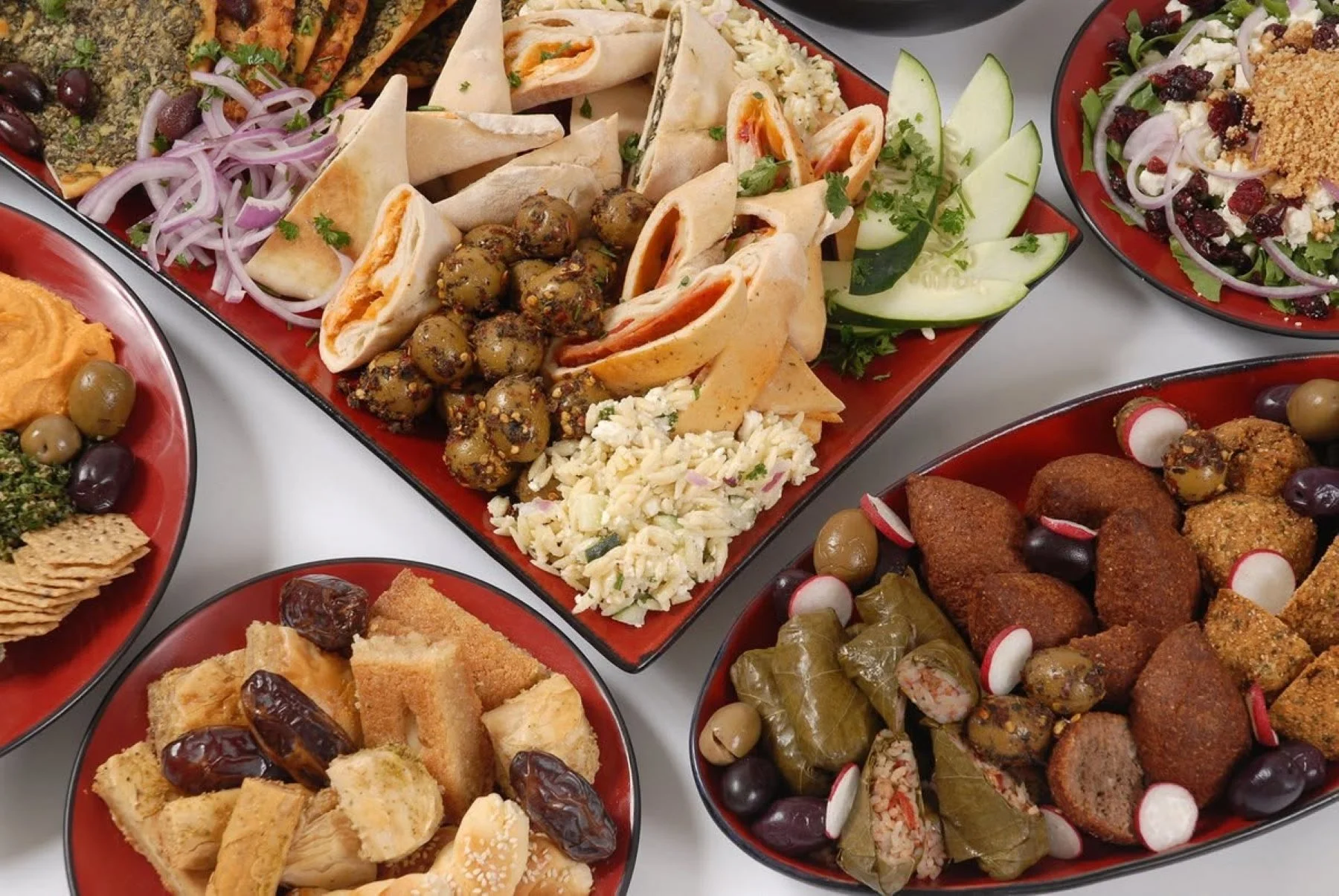 Various Middle Eastern dishes on red plates, including stuffed grape leaves, kibbeh, tabbouleh, falafel, salads, and pita bread with accompaniments.