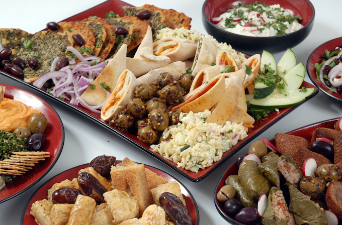 Overhead view of a platter with falafel, pita bread, cucumber slices, and various side dishes including yogurt sauce and tabbouleh, surrounded by smaller plates of stuffed grape leaves, olives, and other Mediterranean appetizers.