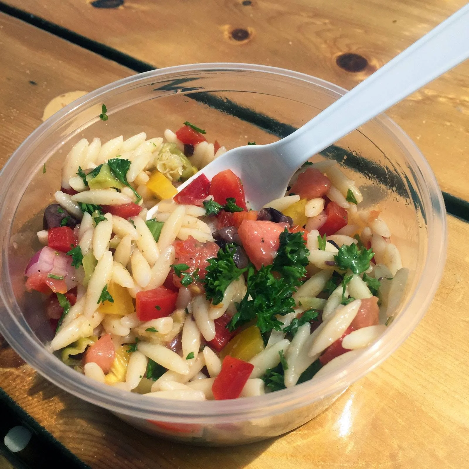A plastic container filled with pasta salad containing cherry tomatoes, chopped onions, parsley, and possibly some chopped peppers, with a white plastic fork inside, resting on a wooden surface.