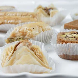 Assorted baklava pastries in white cupcake liners with flaky, nut-filled, and syrupy layers on a white surface.