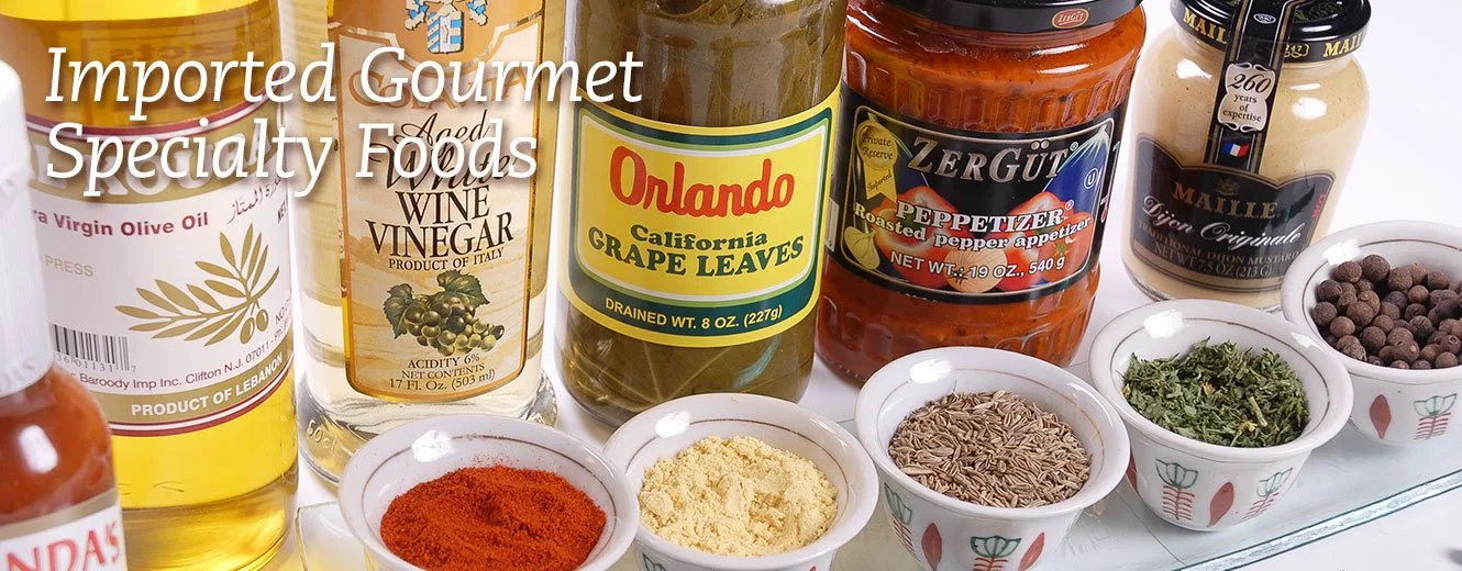 Assorted imported gourmet specialty foods, including vinegar, grape leaves, roasted pepper, mustard, dried herbs, seeds, and spices on a white surface.