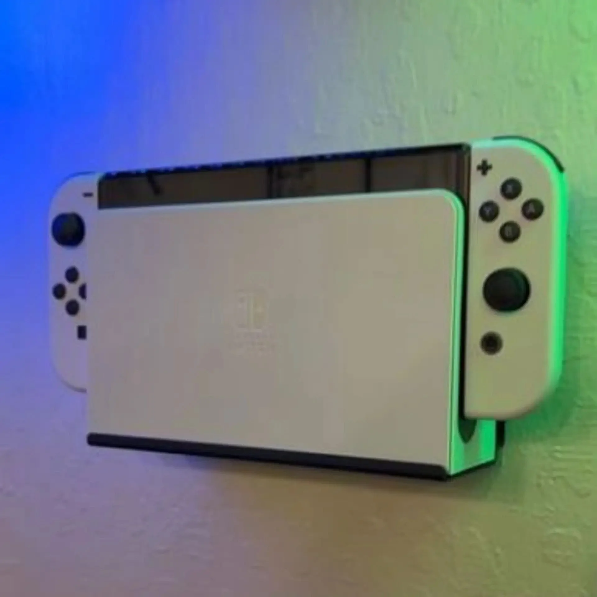 A white gaming console with two controllers, one of them with neon green lighting, placed on a surface with a colorful LED light background. Wall mounting