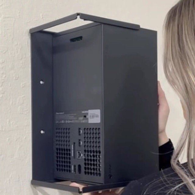 Person wall mounting a black electronic device or box, seen from the side, with a white textured wall in the background.