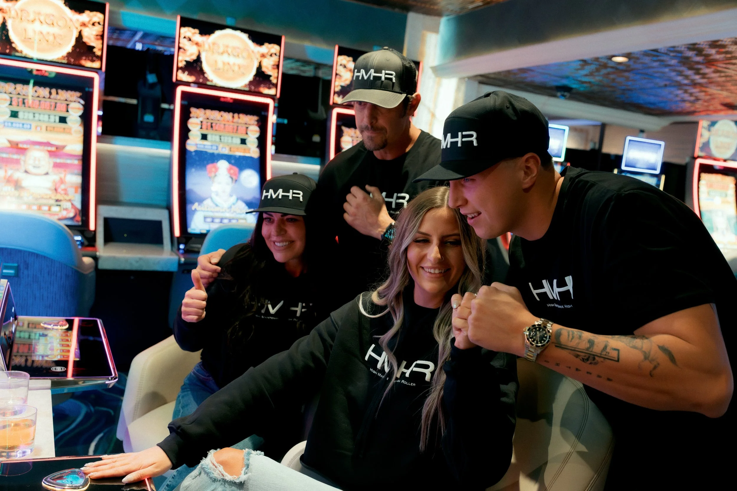 Group of four young adults, two men and two woman, inside a casino, smiling and playing a game at a table with slot machines in the background.