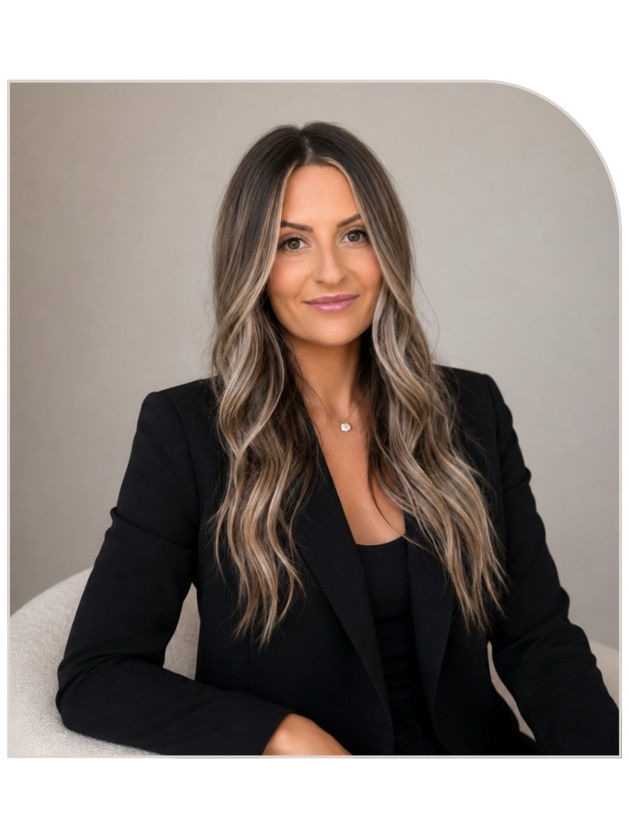 A woman with long wavy hair wearing a black blazer and top, sitting on a light-colored chair against a plain background, smiling at the camera.