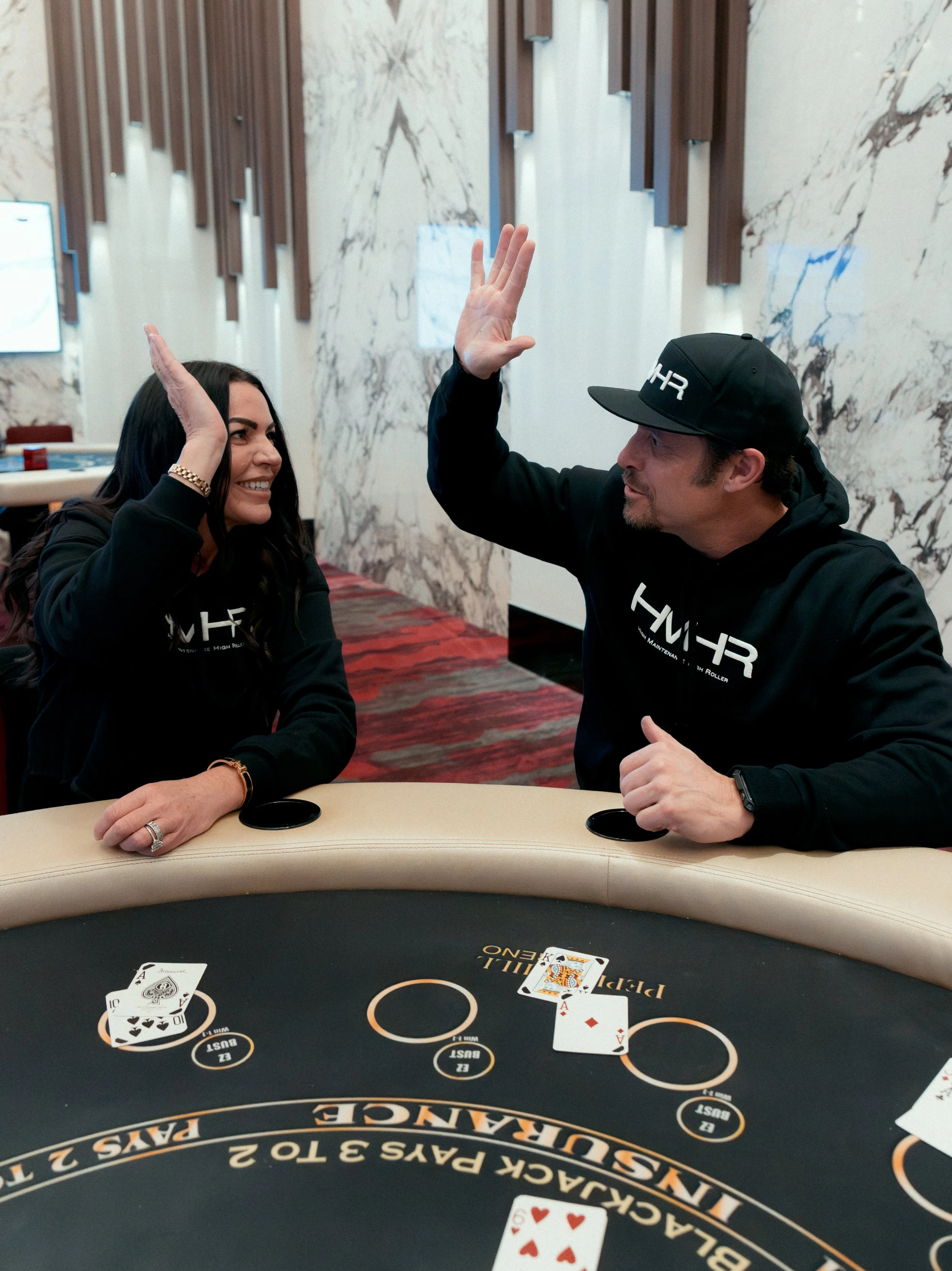 Two people playing poker at a casino table. They are giving each other a high five. The poker table has playing cards and chips. The background features a marble wall with wooden accents.