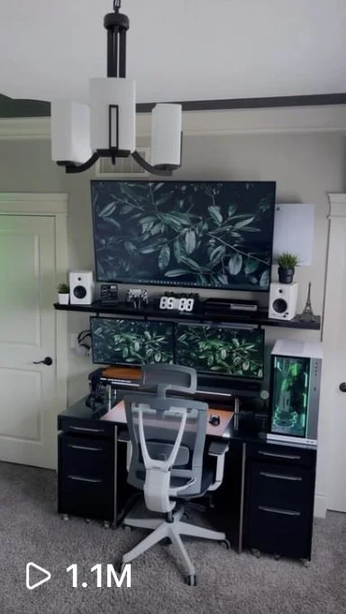 Home office setup with a desk, ergonomic chair, large monitor displaying greenery, and computer tower, in a room with white walls and ceiling light.