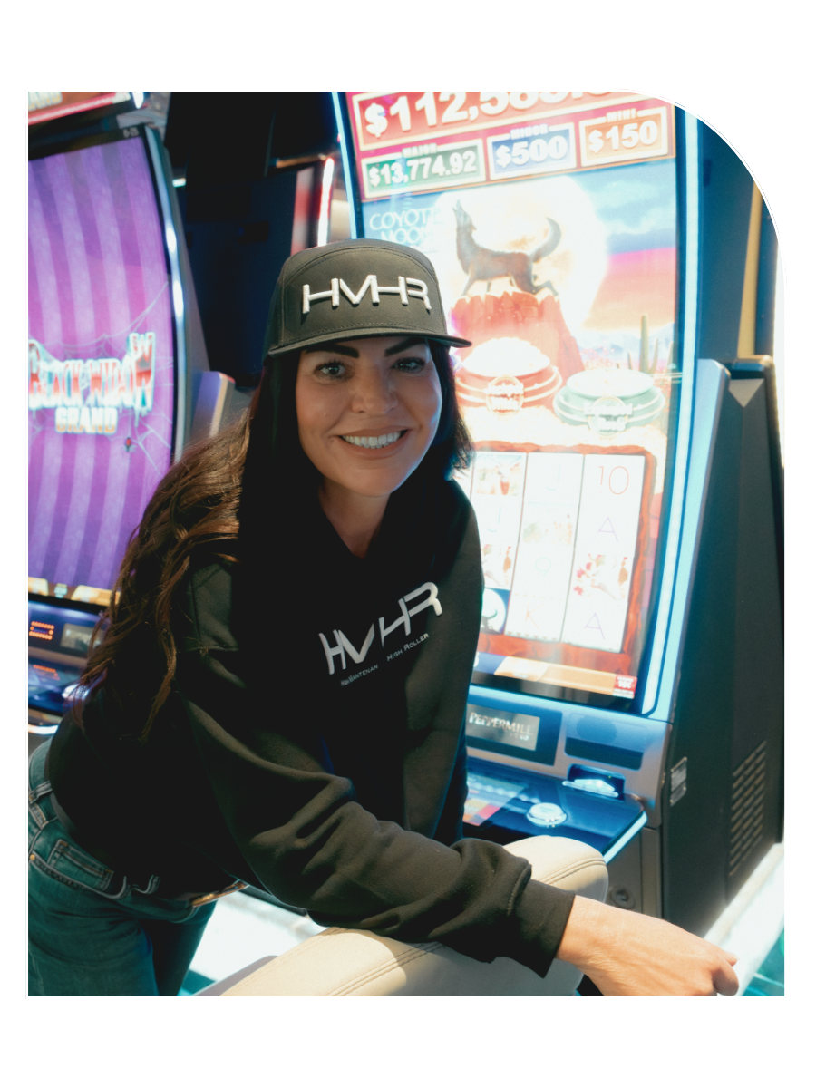 A woman in a black hoodie and cap with 'HMHR' logo, smiling, in front of a casino slot machine displaying a game with a coyote, cactus, and rock formation.