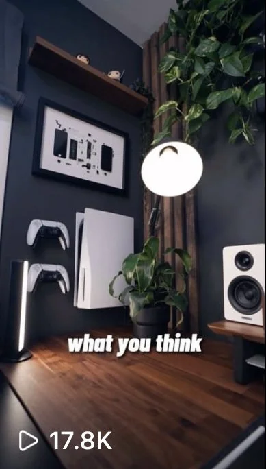 A home office or entertainment setup with a wall-mounted PlayStation 5, a framed art piece, a white speaker, a desk lamp, a wooden desk, and a large green plant next to a window with curtain.