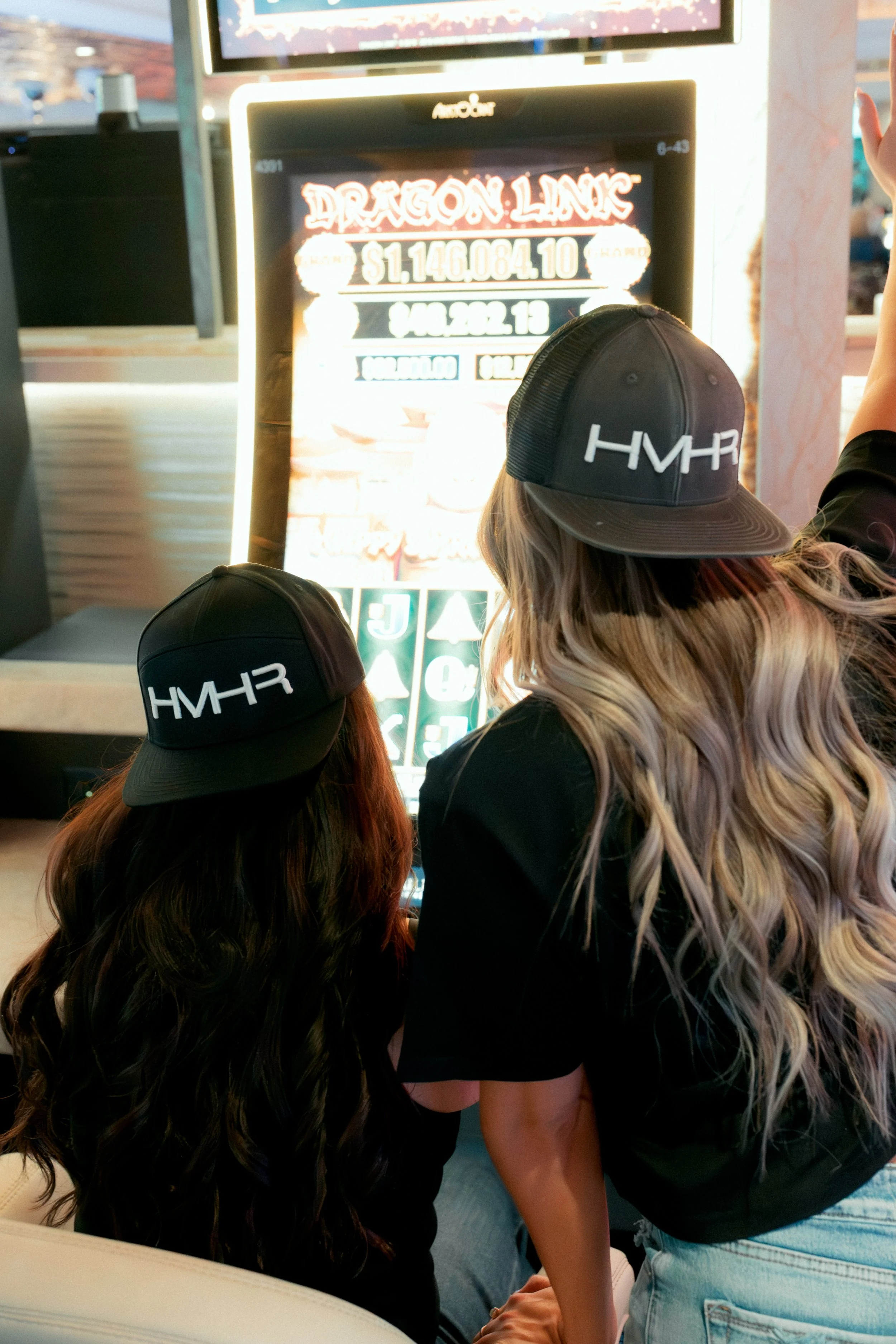 Two women with long, curly hair wearing black hats with the word 'HVR' on them, standing in front of an electronic game display with the title 'Dragon Liar,' at an indoor arcade or amusement venue.