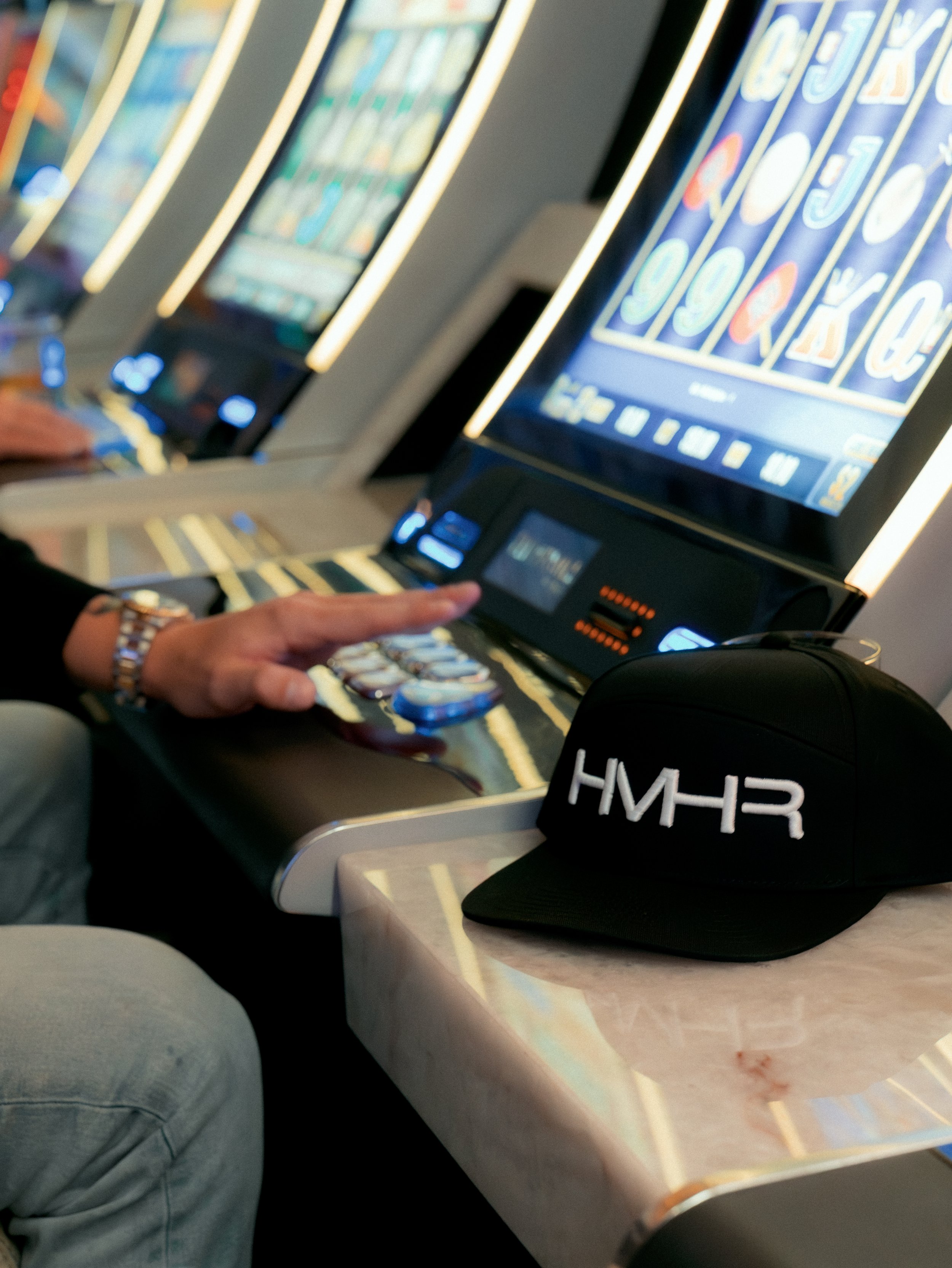 Close-up of a person's hand and arm playing a slot machine in a casino, with a black cap with white text 'HMR' placed on the table in front of the person.