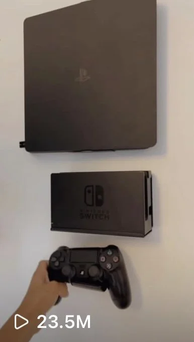 A PlayStation 4 console mounted on a wall with a Nintendo Switch dock and a PlayStation controller being held in a hand below.