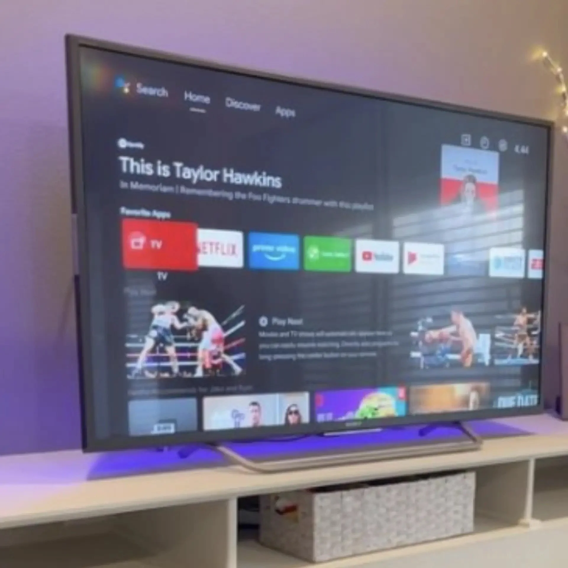 A flat-screen television displaying a streaming service menu with various app icons, including Netflix, YouTube, and Disney Plus, on a white entertainment stand with open shelves below.
