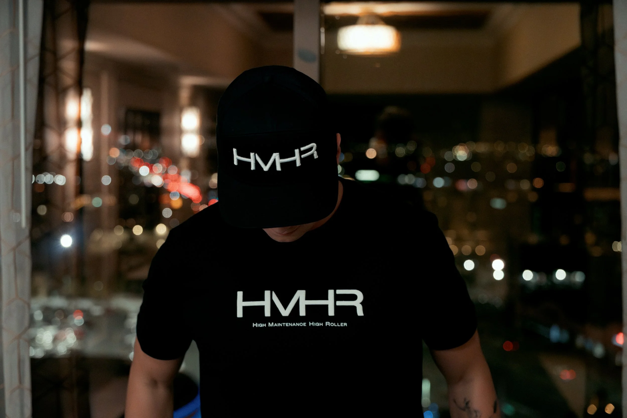 Person wearing a black hoodie and black cap with white text, standing on a balcony with a blurred cityscape in the background at night.
