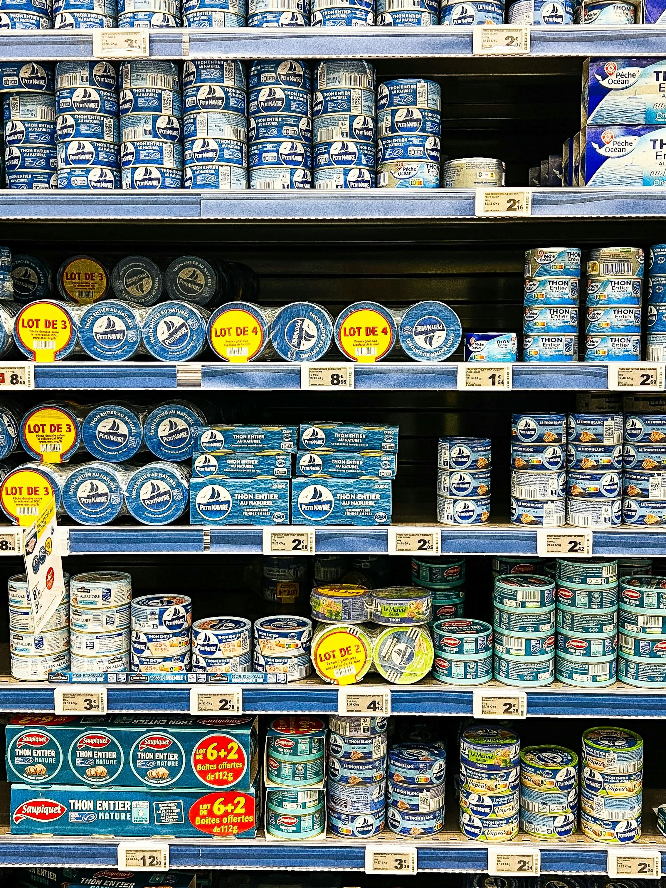 Shelves stocked with cans of tuna fish in a grocery store, with various brands and packaging styles visible.