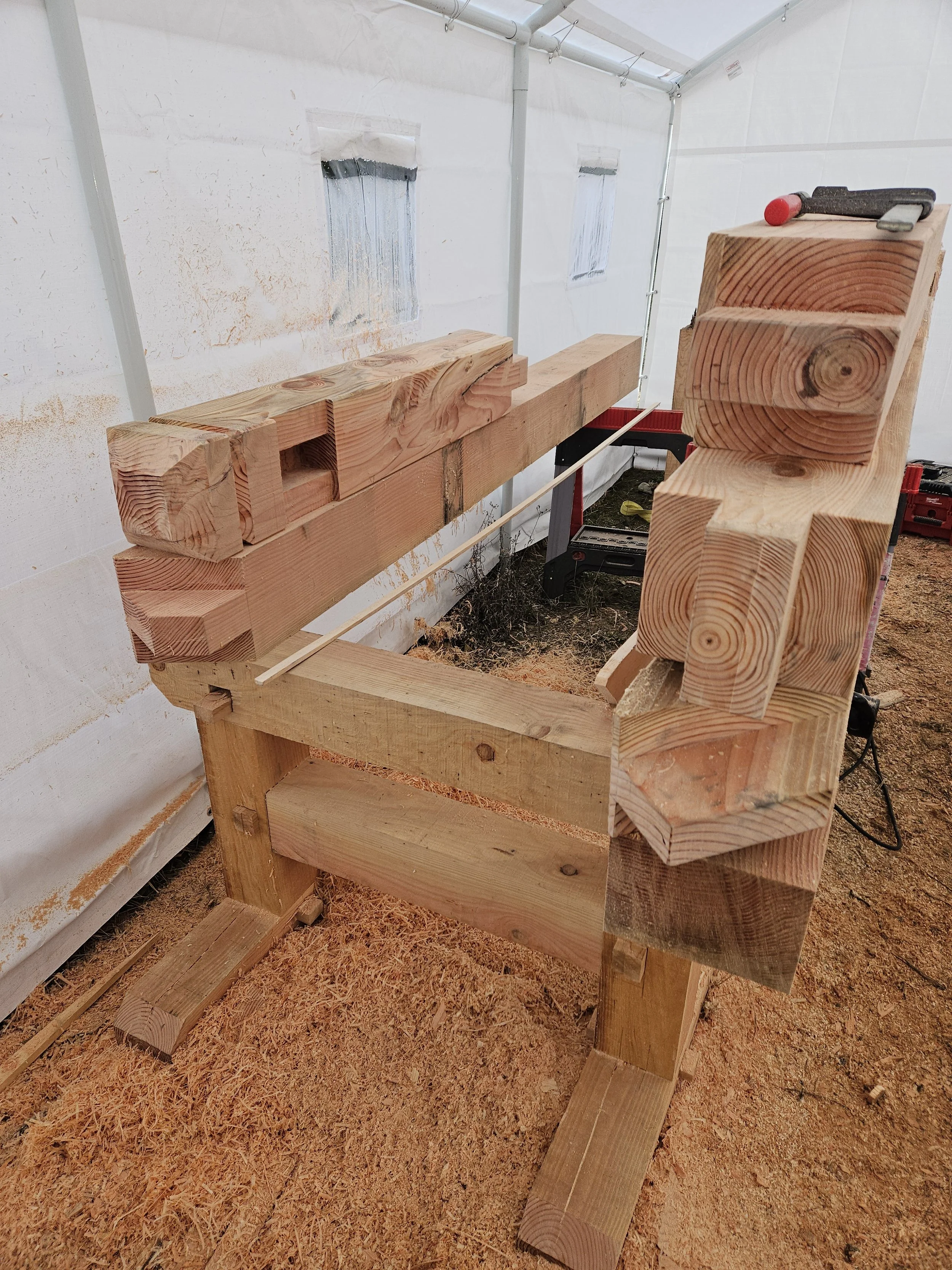 A woodworking workbench inside a white temporary shelter with wood shavings on the ground and various wooden parts and tools on the bench.