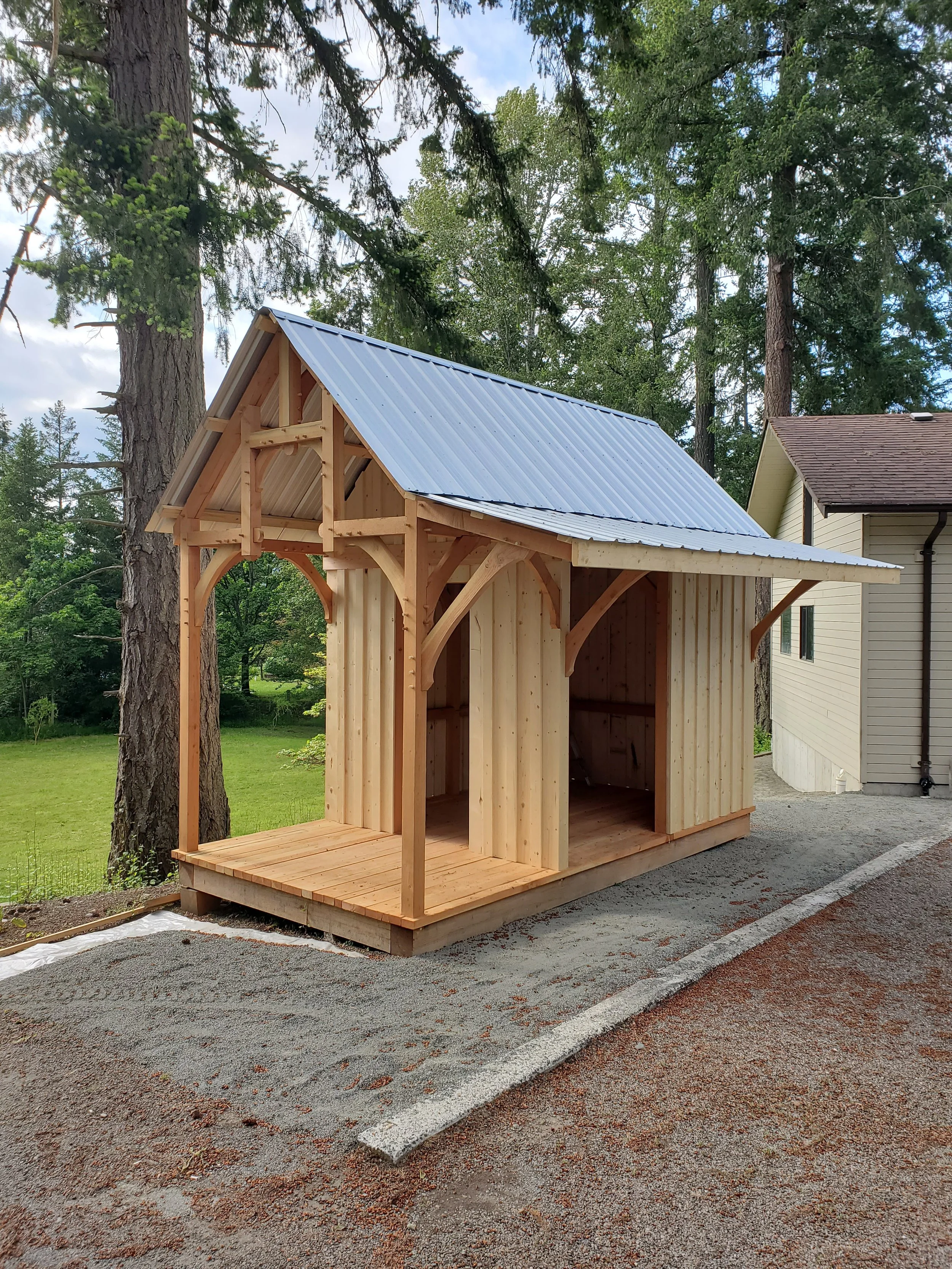 A small wooden structure under construction next to a house, with a metal roof and decorative wood features, surrounded by tall trees and a gravel driveway.