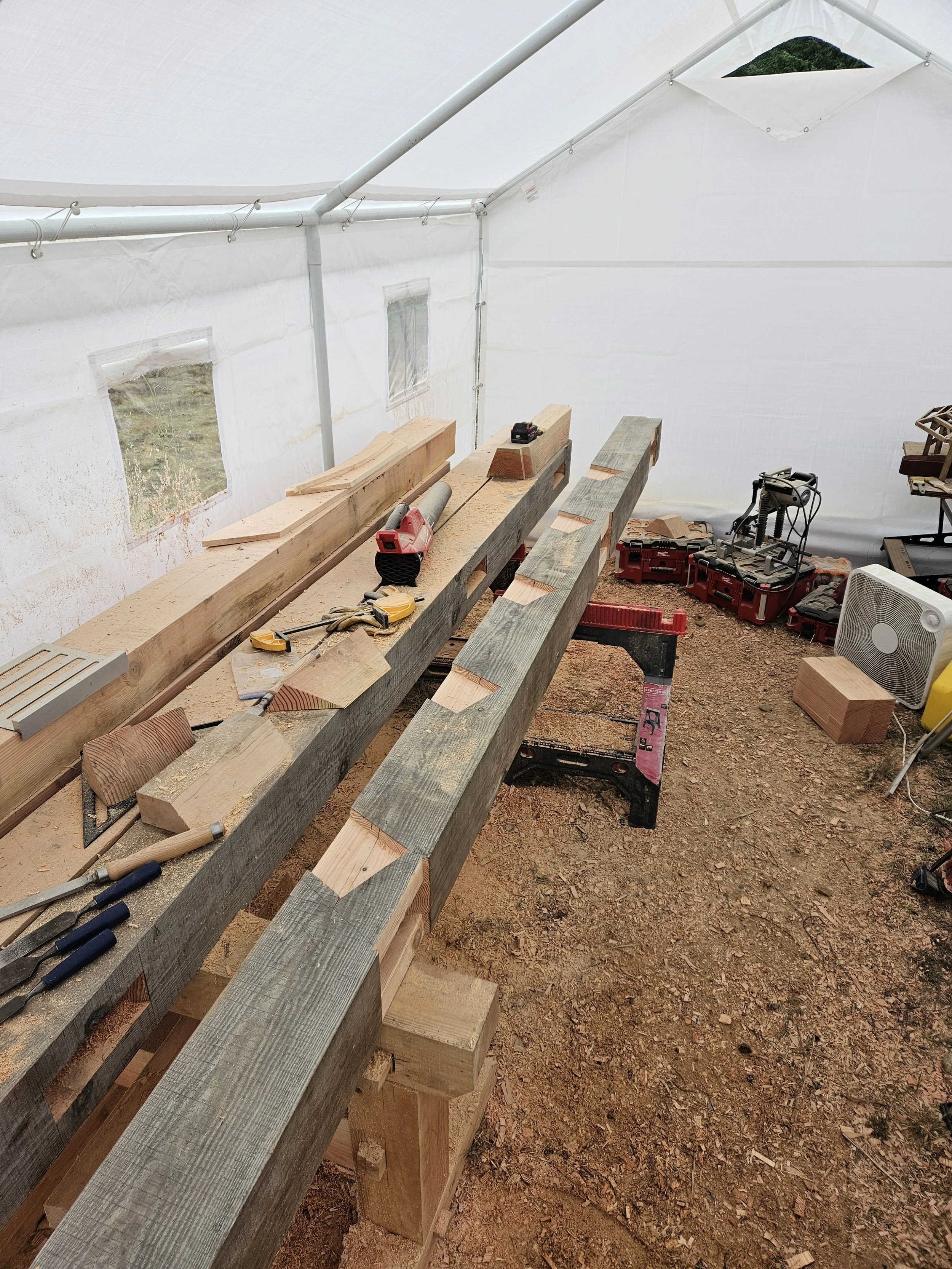 Inside a white tent, wooden planks and construction tools are laid out on workbenches. There are windows in the tent walls, and various power tools and equipment are visible in the background on the ground.