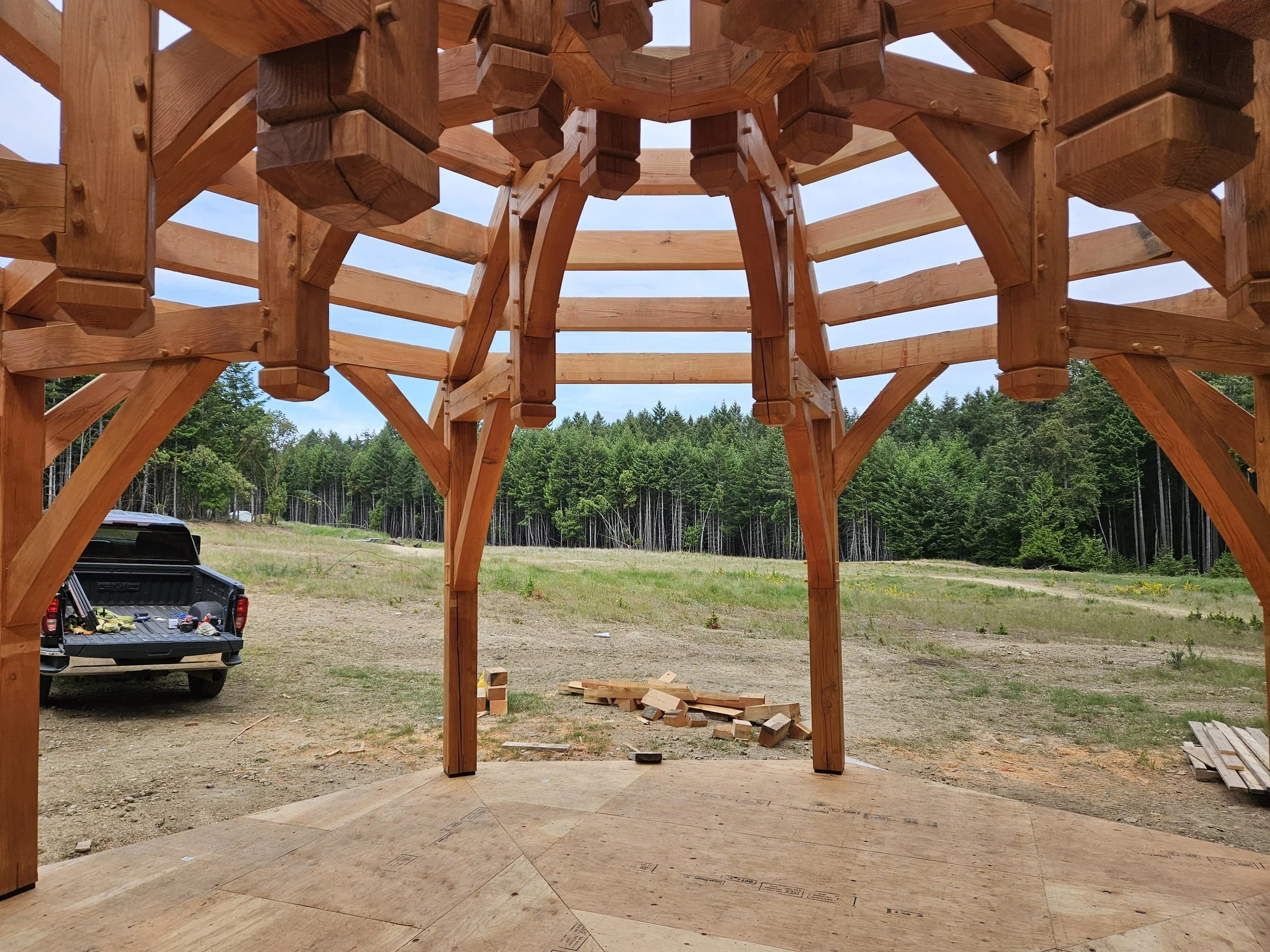 Wooden gazebo under construction with a view of the blue sky, green trees, and a car parked in the background.