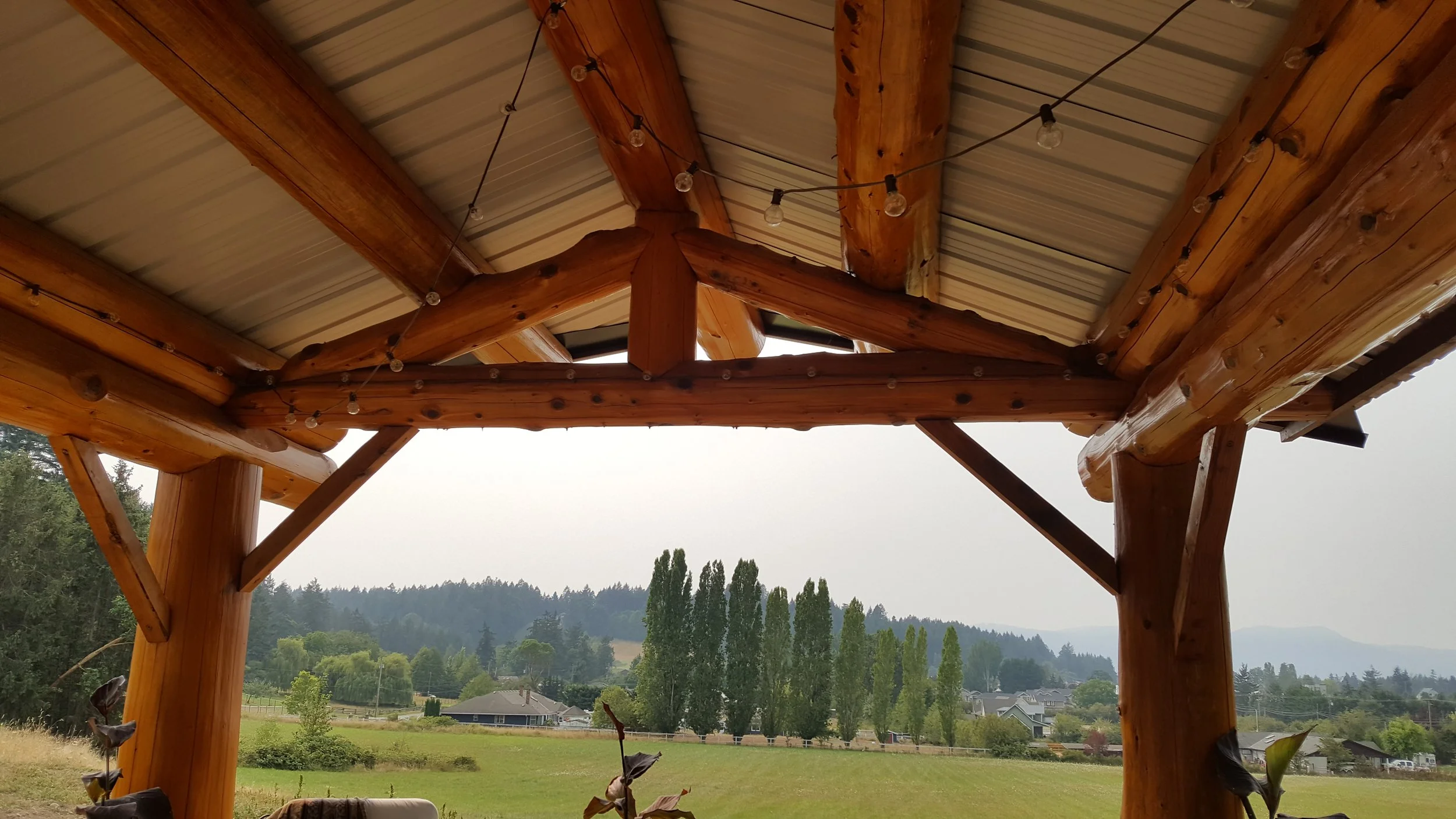 A wooden outdoor structure with a glazed roof, decorated with string lights, overlooking a green landscape with trees and houses in the distance.