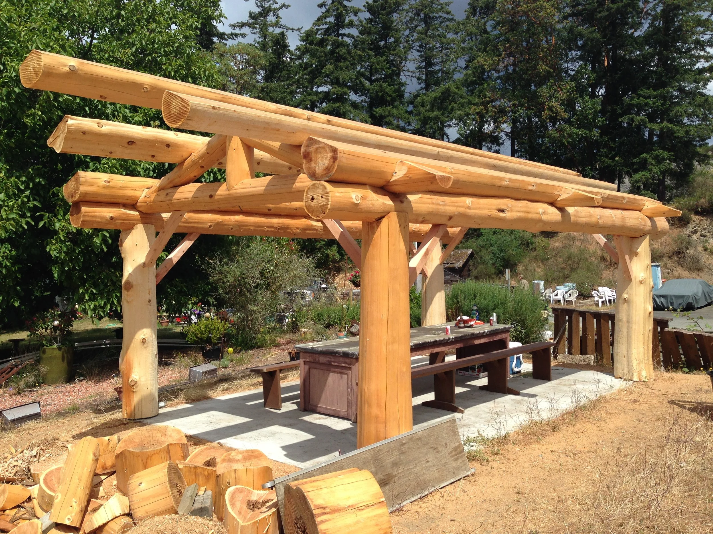 Wooden beams and logs for a construction project, with some finished and some in progress, outdoors with trees and a seating area in the background.