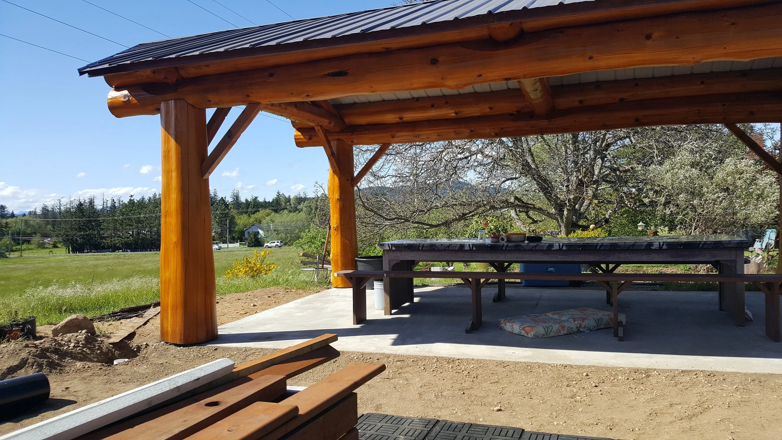 Wooden outdoor pavilion with a roof, supported by large wooden beams, overlooking a grassy yard with trees, a bench, and concrete patio area.