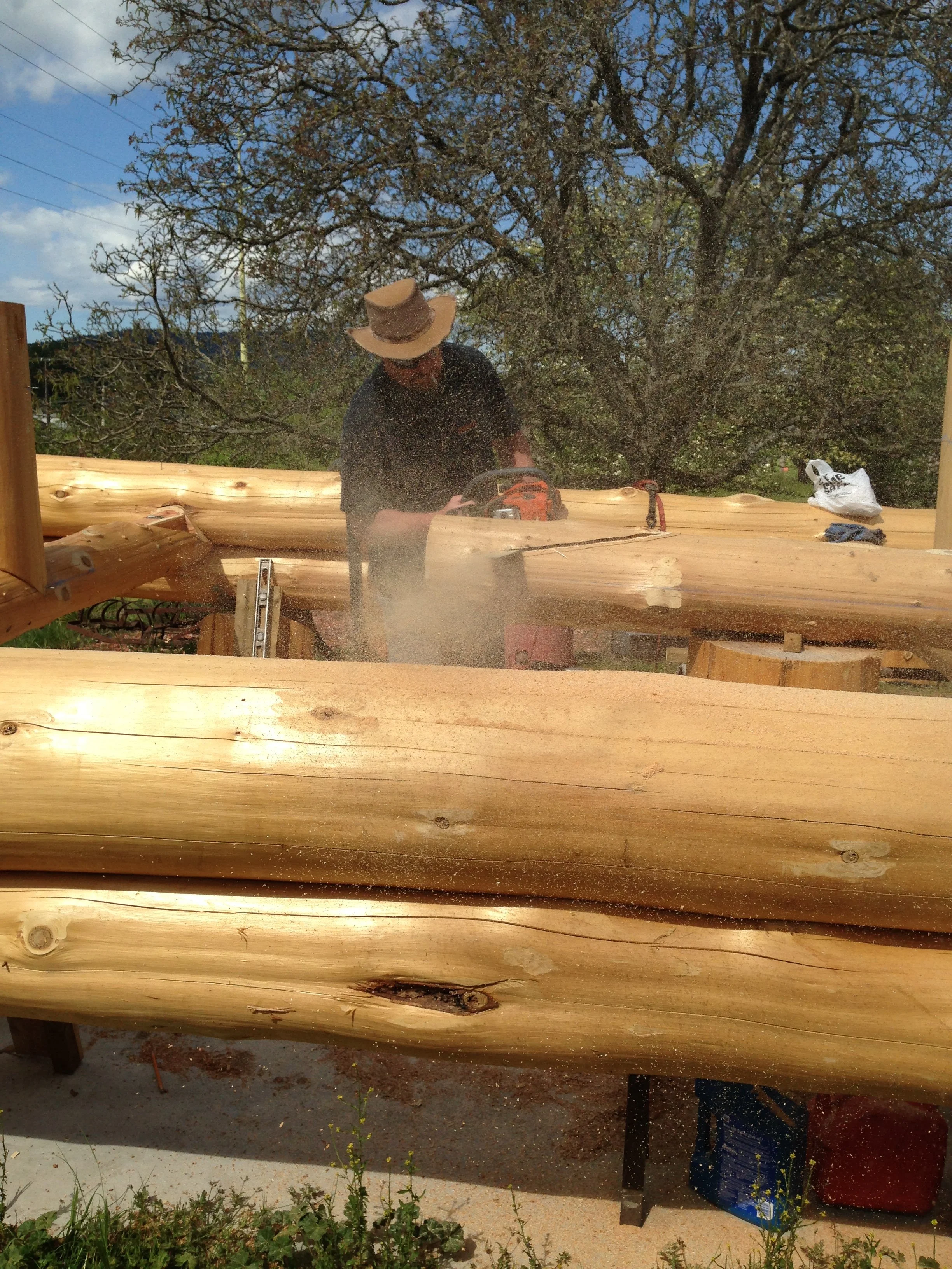 A person wearing a hat and black shirt using a chainsaw to cut a large log of wood outdoors with trees and a partly cloudy sky in the background. There are tools and a bag on the wood surface.